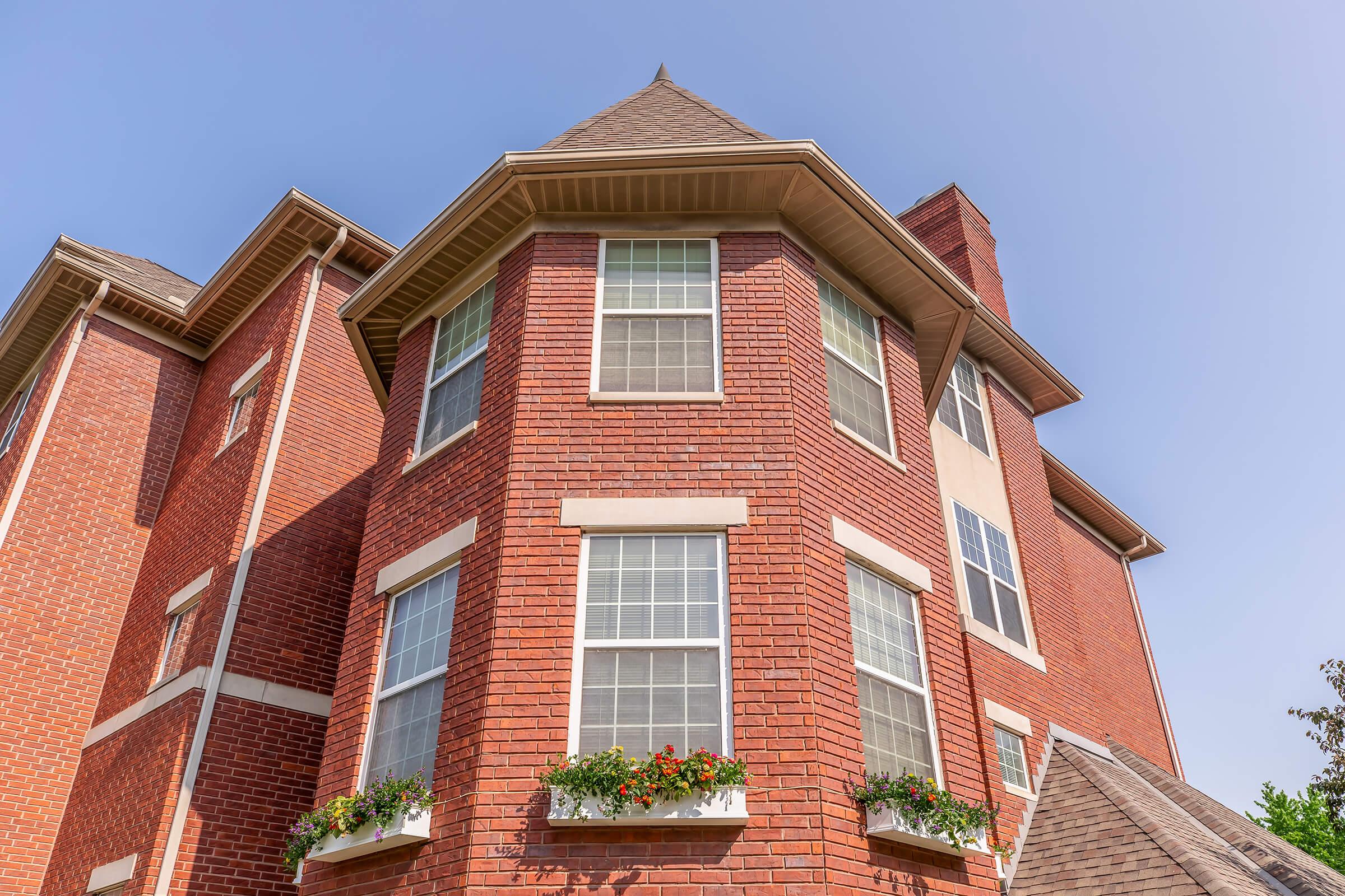A close-up view of a brick house featuring large windows and flower boxes. The building has a triangular roof and is set against a clear blue sky. The angle captures the architectural details and textures of the red brick exterior.