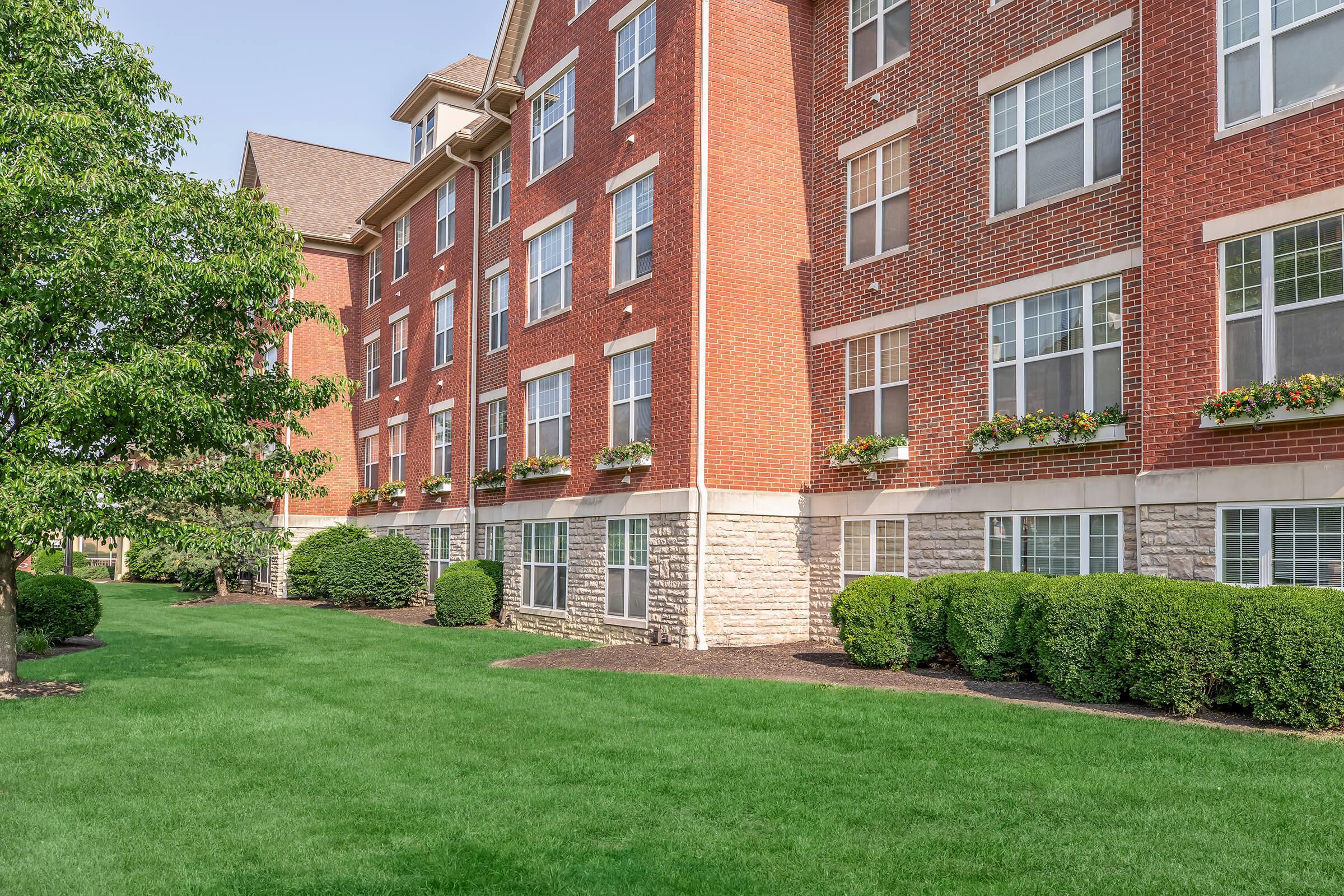 A brick apartment building featuring multiple floors with large windows. The exterior includes decorative flower boxes under the windows and well-maintained greenery, including neatly trimmed bushes and a lawn, creating a welcoming environment.
