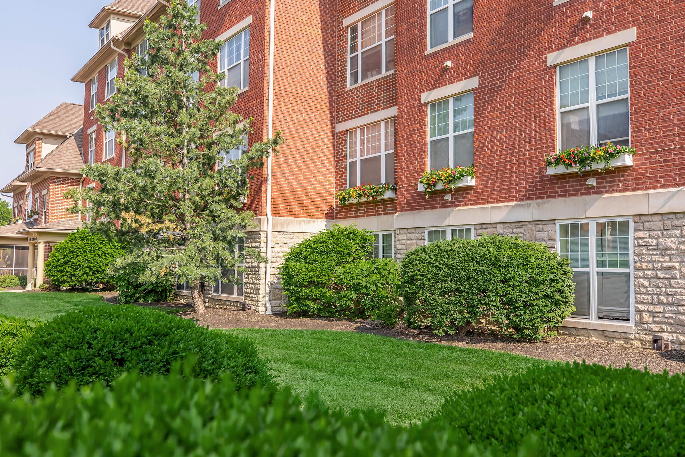 A well-maintained exterior view of a red brick building with large windows, potted flowers on the ledges, and neatly trimmed greenery in the foreground. The landscaping includes lush green bushes and a manicured lawn, creating a welcoming atmosphere.