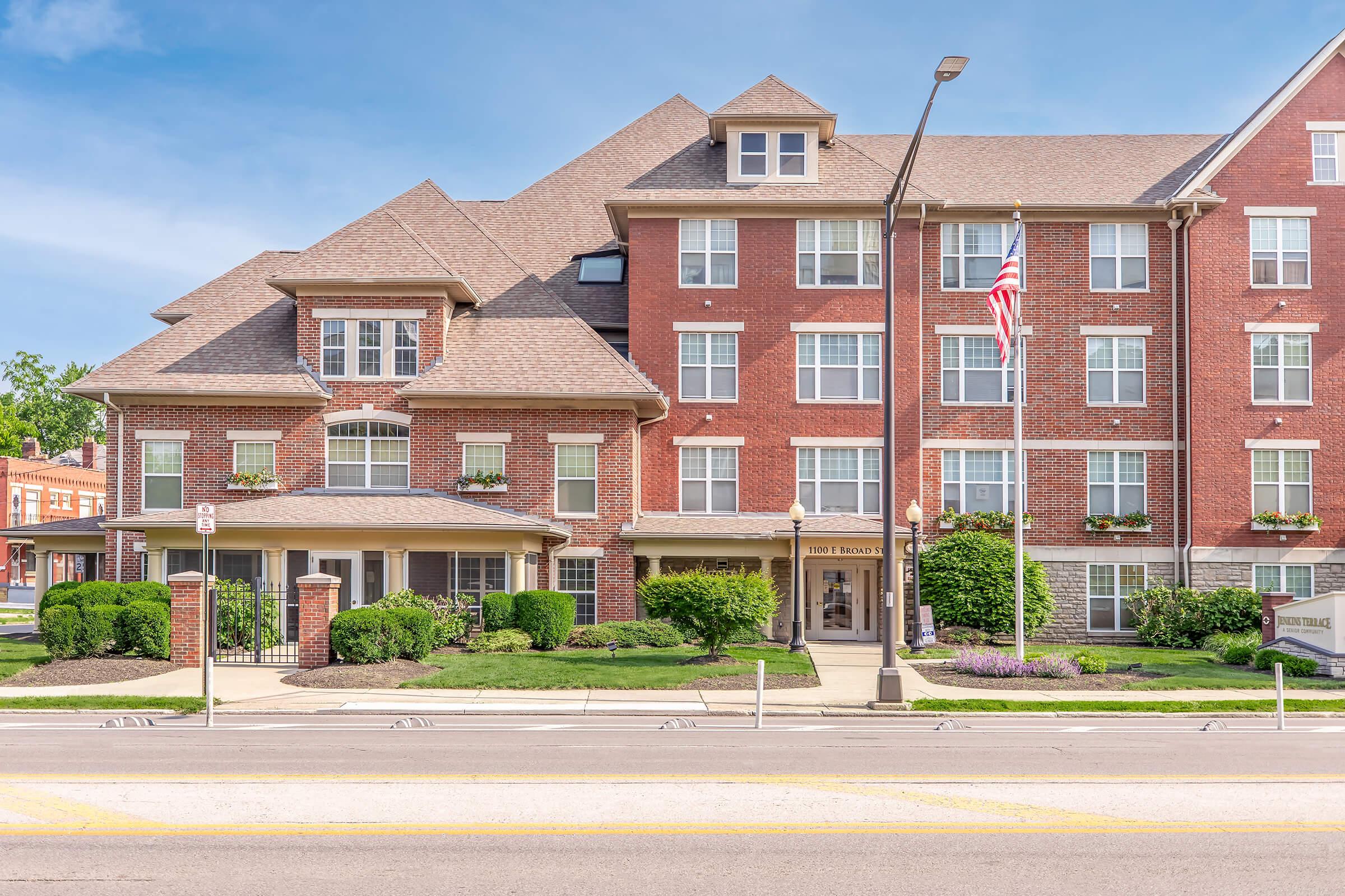 A brick apartment building with a peaked roof, featuring multiple windows and flower boxes. An American flag is displayed outside. The entrance has a covered porch with decorative landscaping, and the street in front is visible, indicating an urban setting.