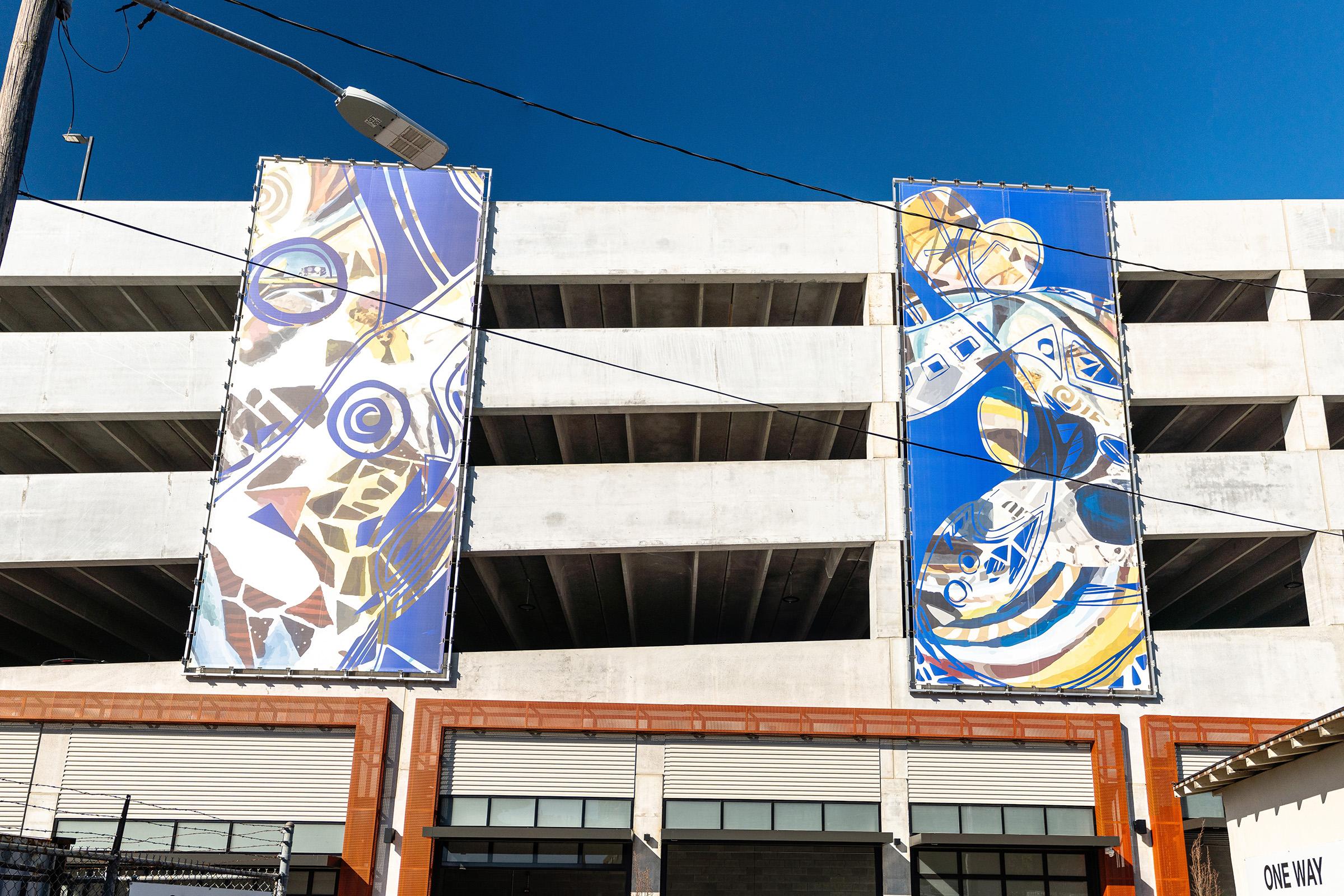 Two large, colorful murals are displayed on the side of a concrete parking garage. The murals feature abstract designs with vibrant blue, gold, and white elements. Power lines cross in front, and the lower part of the building is partially obscured by a fence and commercial storefronts below.