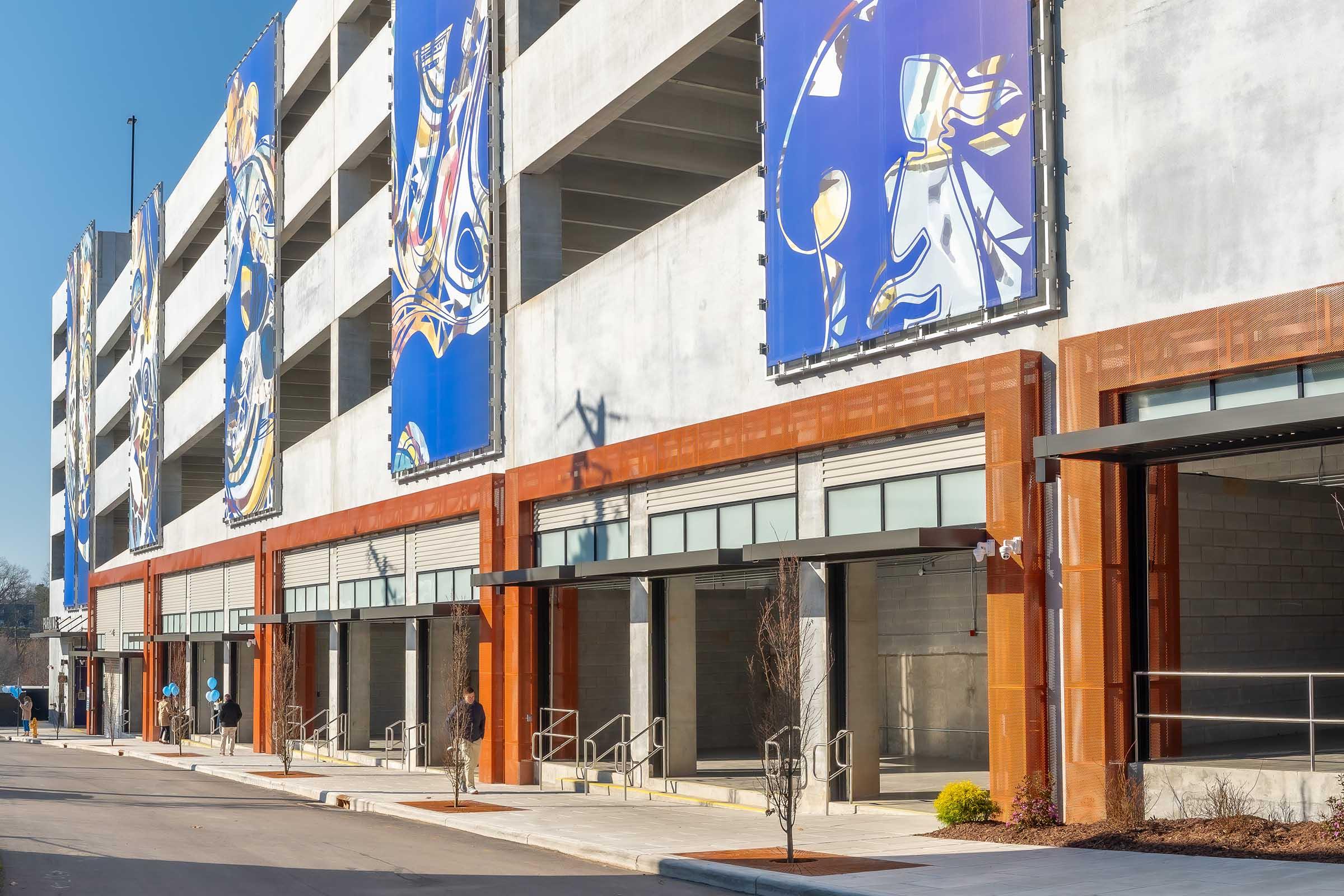 A modern multi-story parking garage with large blue banners and rust-colored metal accents. The building features a clean design with prominent loading areas and modern architectural elements. The surrounding area includes paved pathways and landscaping.