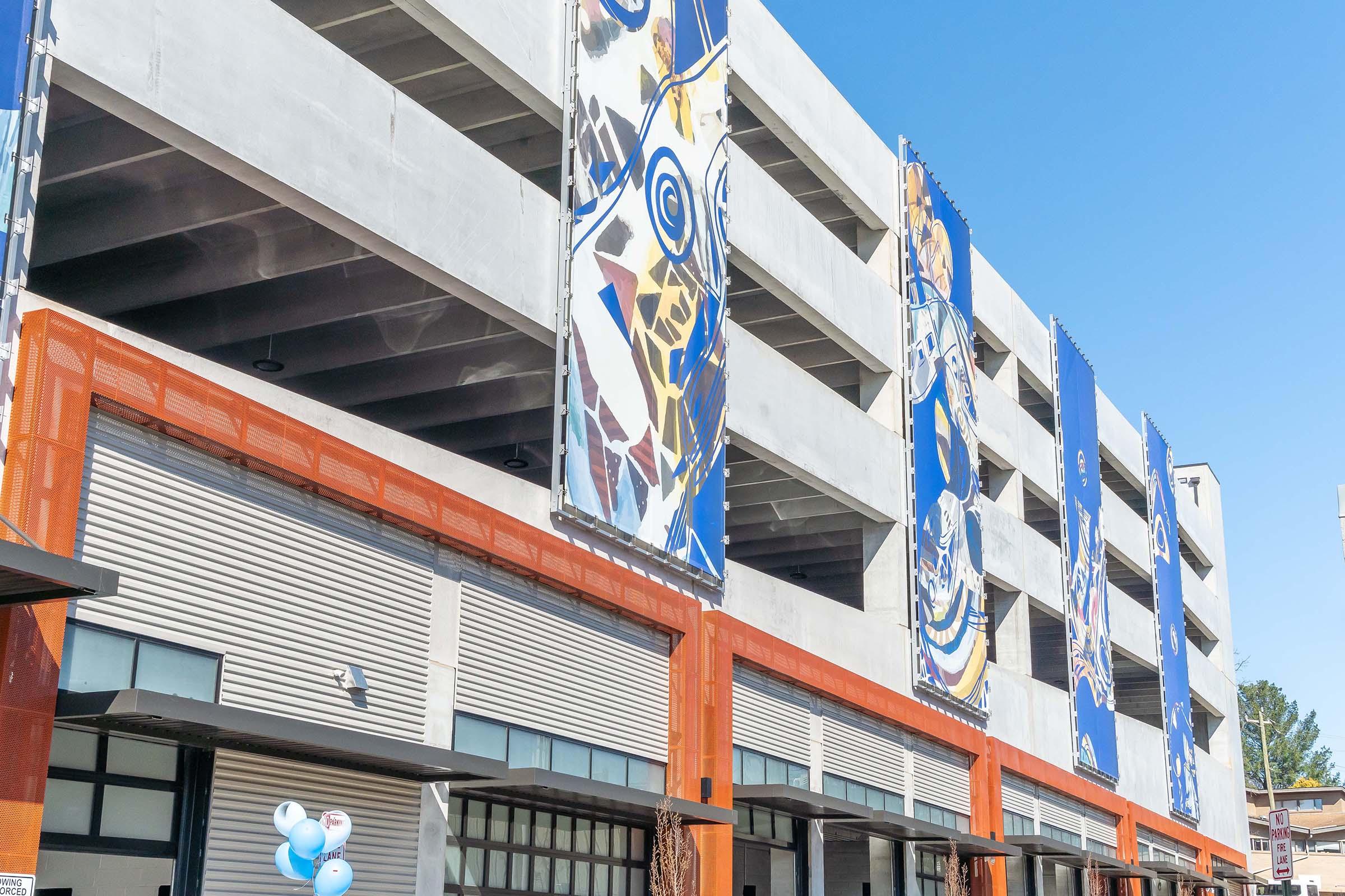A multi-level parking structure featuring large, colorful murals on the exterior walls. The building displays an orange accent at the base and is set against a clear blue sky. Balloons are visible in the foreground, suggesting a celebratory atmosphere.
