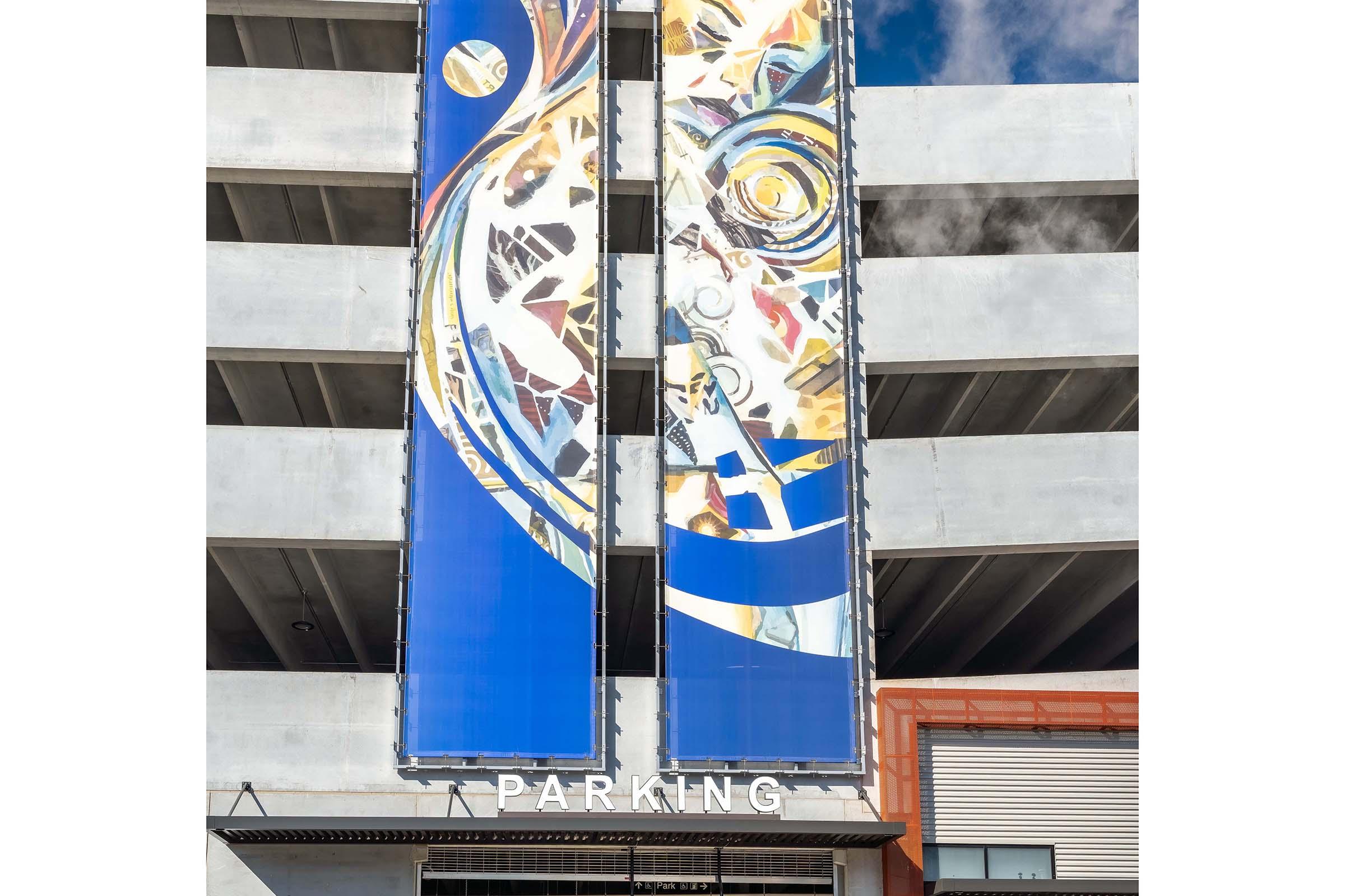 A modern multi-level parking structure featuring a large, colorful mural on two banners. The mural includes abstract designs in blue, yellow, and white. Below the artwork, the word "PARKING" is prominently displayed in bold letters, indicating the building's function. The sky is visible above, adding depth to the scene.