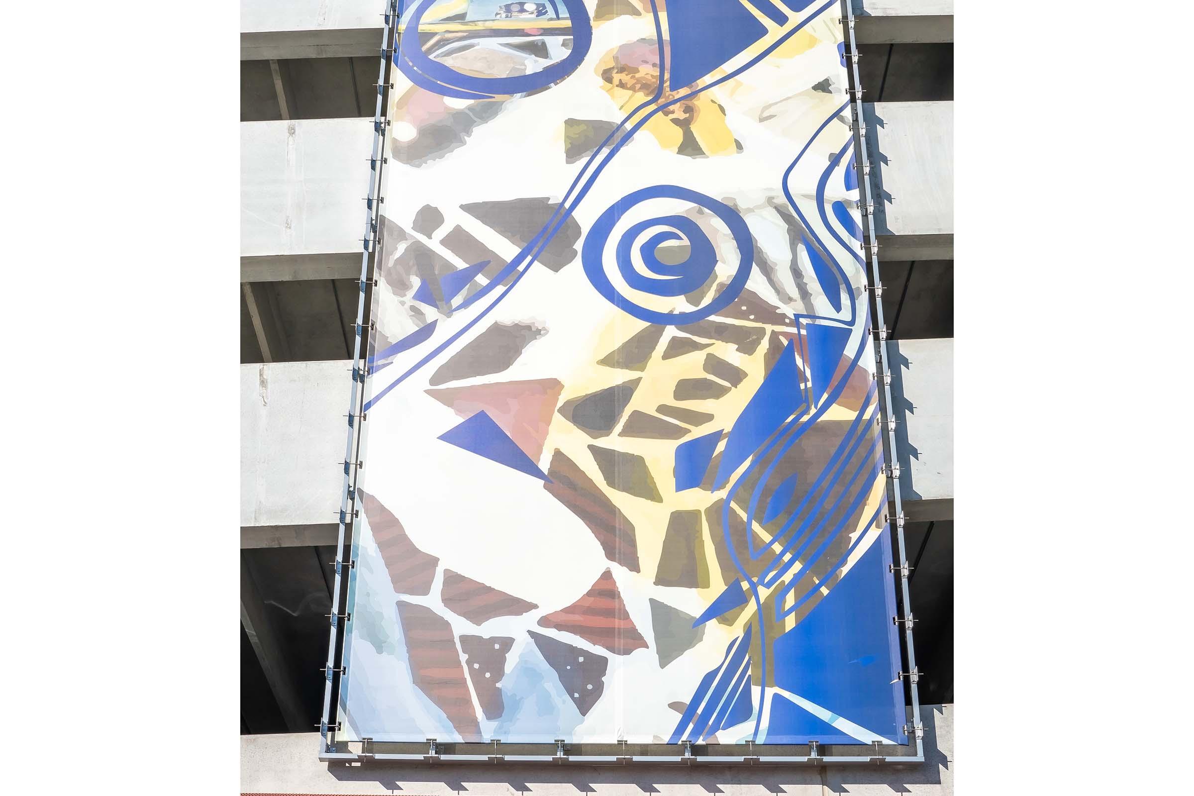 Abstract mural on a concrete parking structure featuring bold colors and shapes, including swirls, geometric patterns, and a mix of blues, yellows, and earthy tones. The mural appears to blend artistic elements with the urban architecture, creating a vibrant visual contrast against the concrete backdrop.