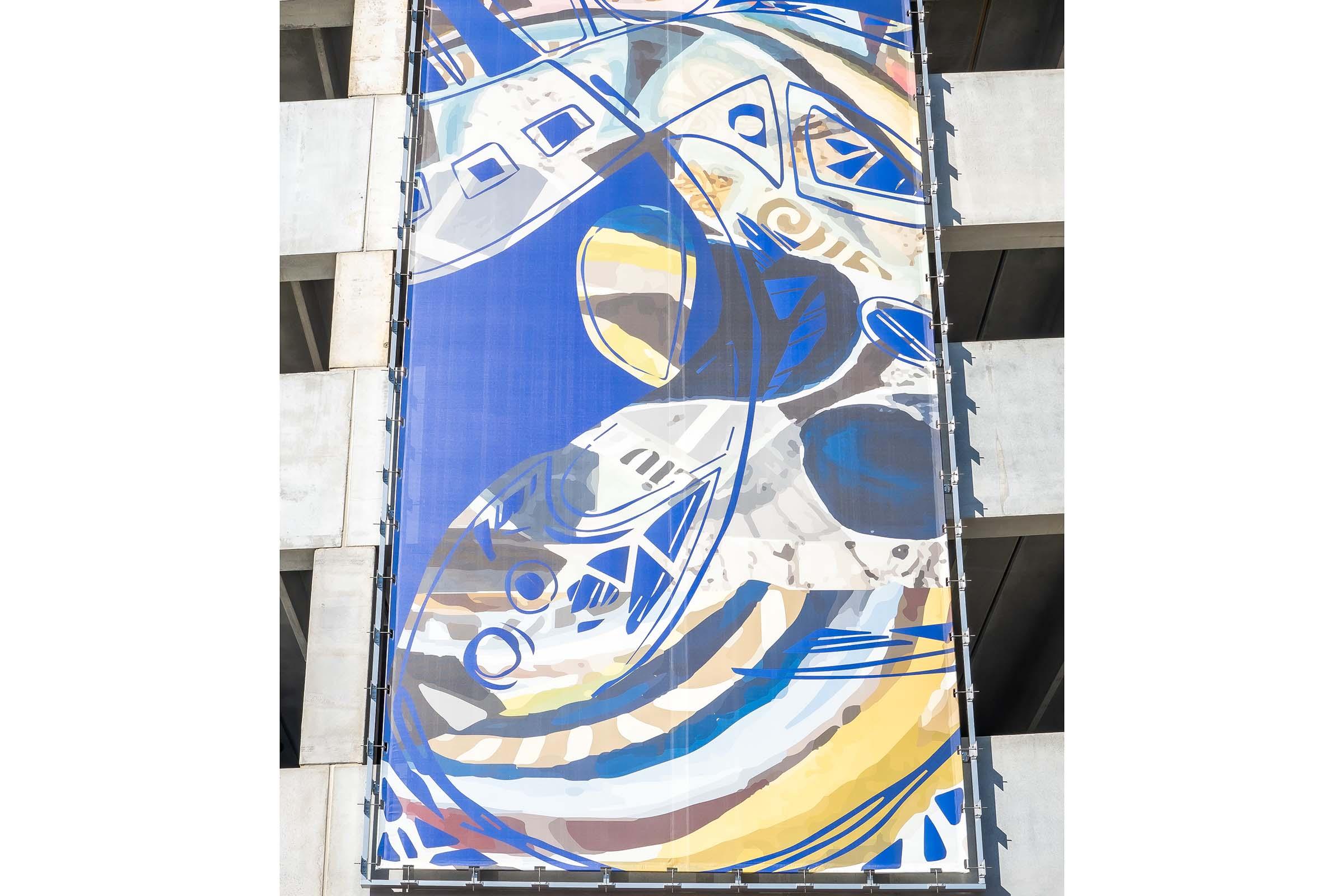 A vibrant mural displayed on a building facade, featuring abstract shapes and colorful designs in shades of blue, yellow, and white, depicting stylized fish and other elements. The artwork adds visual interest to the concrete structure.