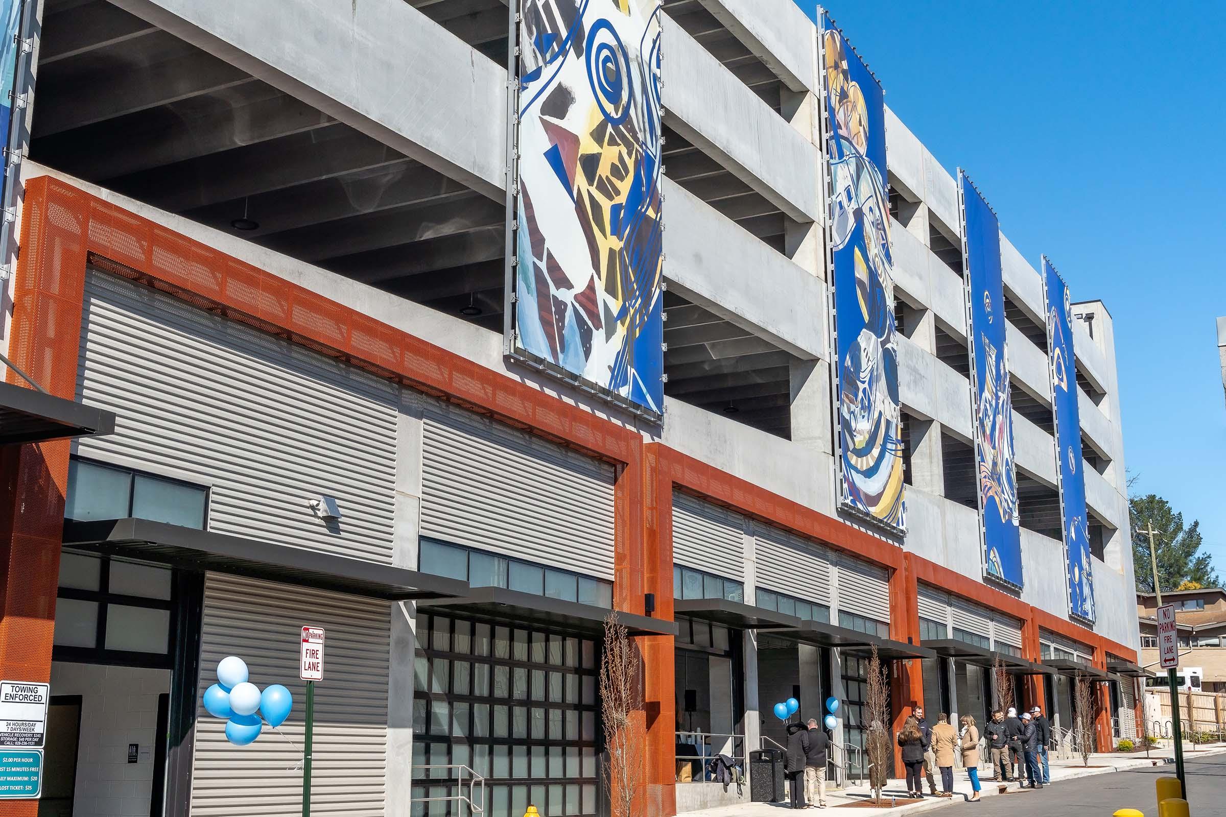 A modern multi-story parking garage with colorful banners displayed on its side. The entrance features a sleek design, and several people are gathered outside, some with blue balloons, on a clear, sunny day. The structure has a mix of concrete and metal elements, showcasing contemporary architecture.