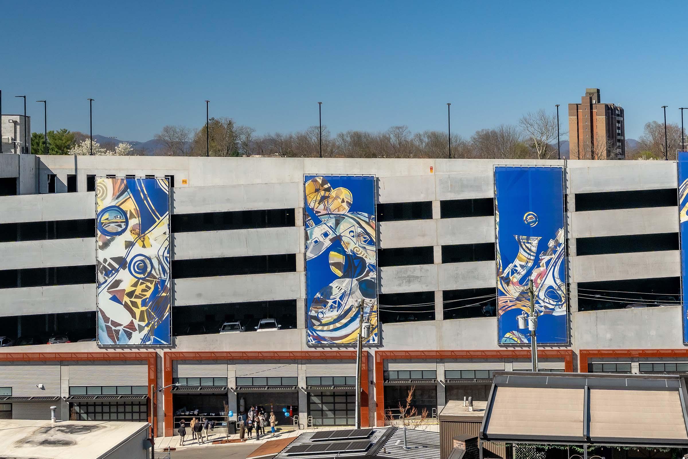 A modern parking garage featuring large, colorful banners with abstract designs. The banners hang vertically on the upper levels, while a busy area below showcases people and outdoor seating. The backdrop includes a clear blue sky and distant trees.
