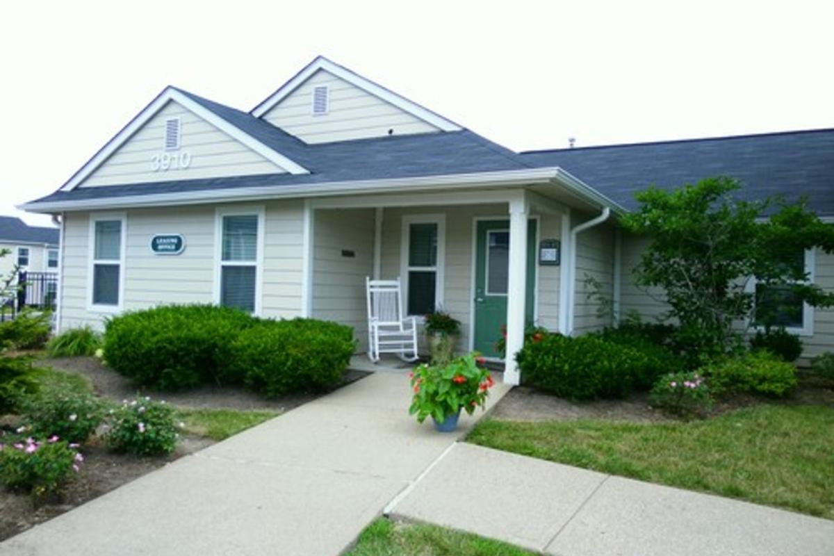 A small, single-story residential building with a porch featuring a rocking chair. The entrance is framed by two large windows, and there are green bushes and flower beds on either side of a concrete pathway leading to the door. The sky is overcast, creating a calm atmosphere.