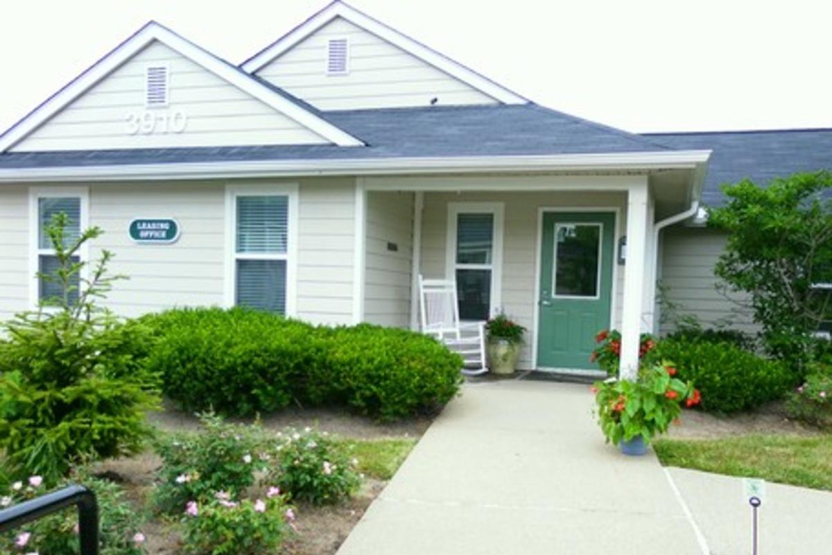 A single-story building with a light-colored exterior and a green door. It features a porch with a rocking chair, surrounded by well-maintained gardens with flowering plants and shrubs. The sign near the door reads "Leasing Office," indicating it serves as an office for rentals.