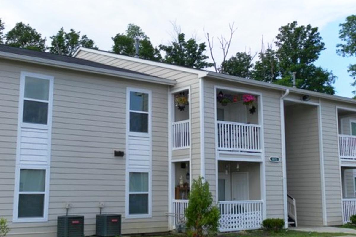 A two-story apartment building with a beige exterior. The building features balconies with hanging flower pots, surrounded by greenery. There are air conditioning units at the base, and a clear sky is visible above.