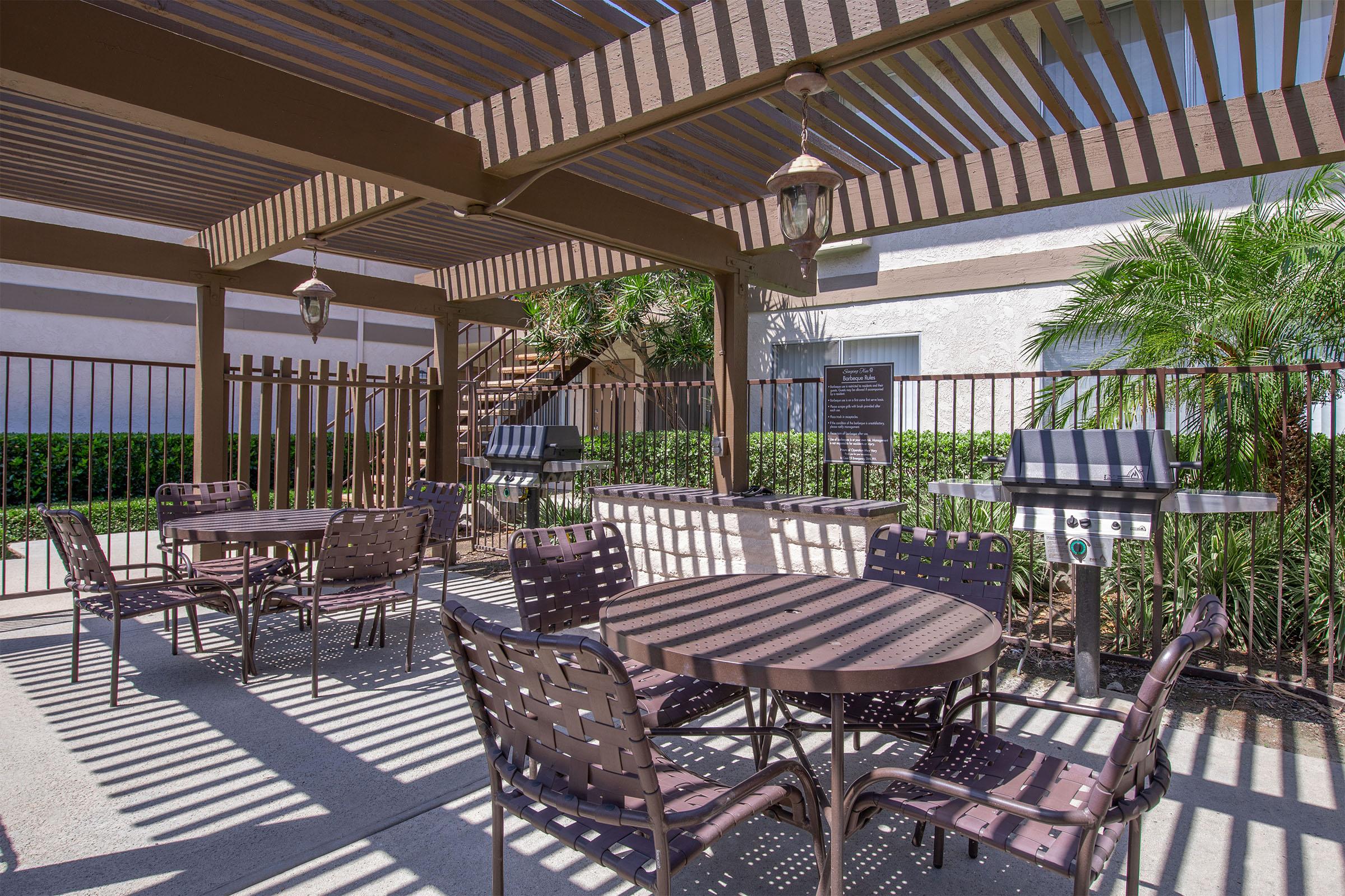A shaded outdoor seating area with several metal tables and chairs under a pergola. There are two barbecue grills nearby, surrounded by lush greenery and a decorative fence. The space is well-lit with natural sunlight filtering through the slatted roof above.