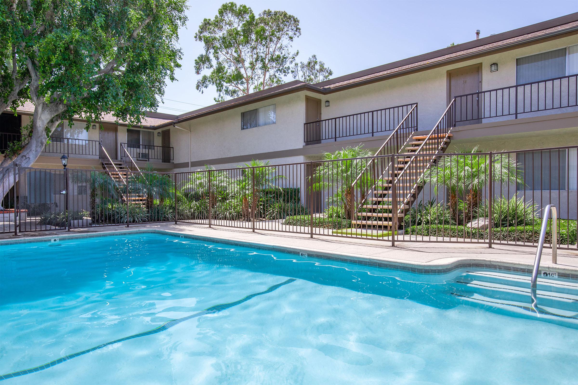 A clear swimming pool in the foreground with sunlight reflecting on the water. In the background, there are two-story apartment buildings with balconies and wooden stairs. Lush greenery, including palm trees and shrubs, surrounds the pool area, creating a relaxing atmosphere.