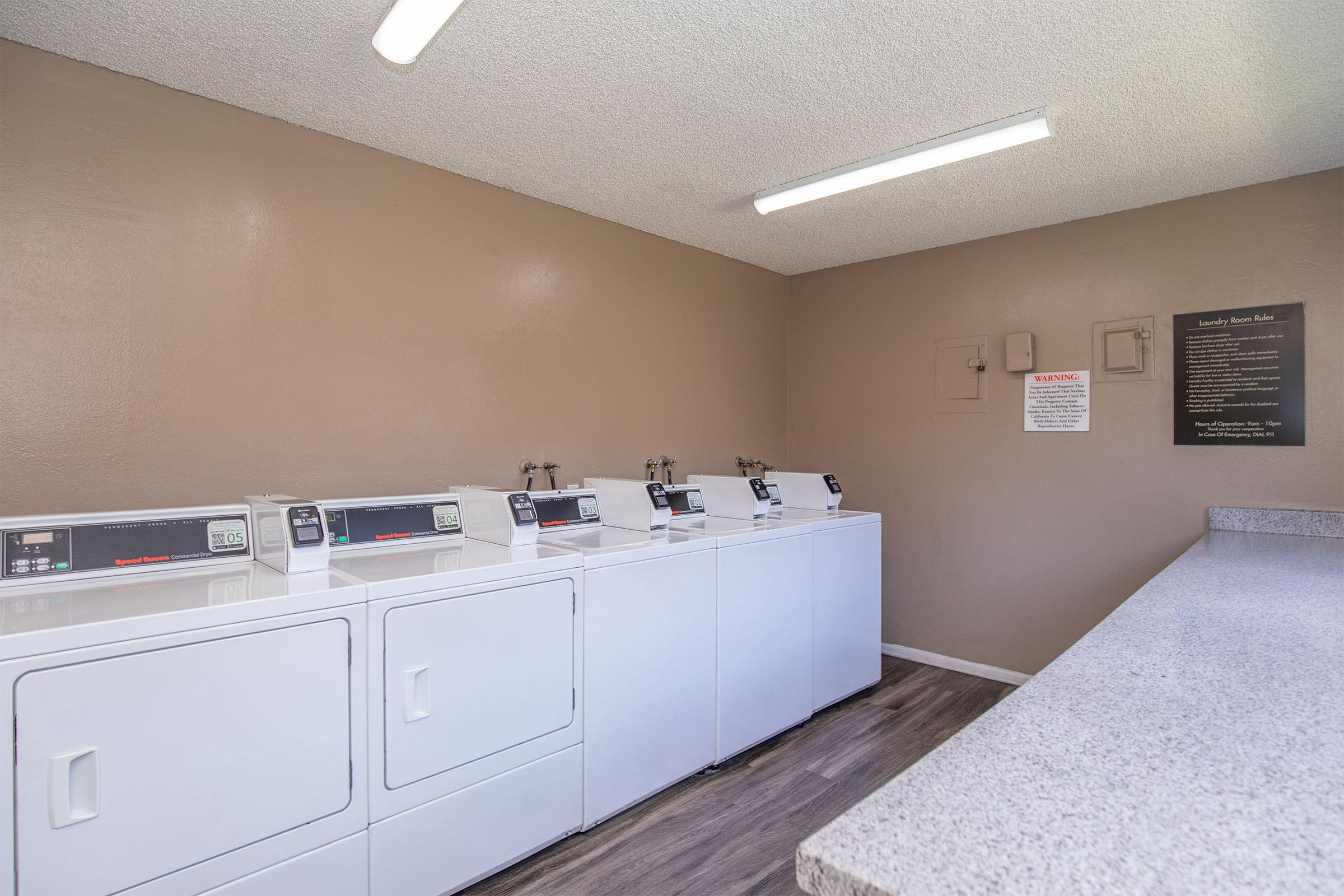 A clean laundry room featuring multiple white washing machines and dryers lined up against a beige wall. There is a countertop for folding laundry and signs detailing laundry room guidelines posted on the wall. The space is well-lit with fluorescent lighting.