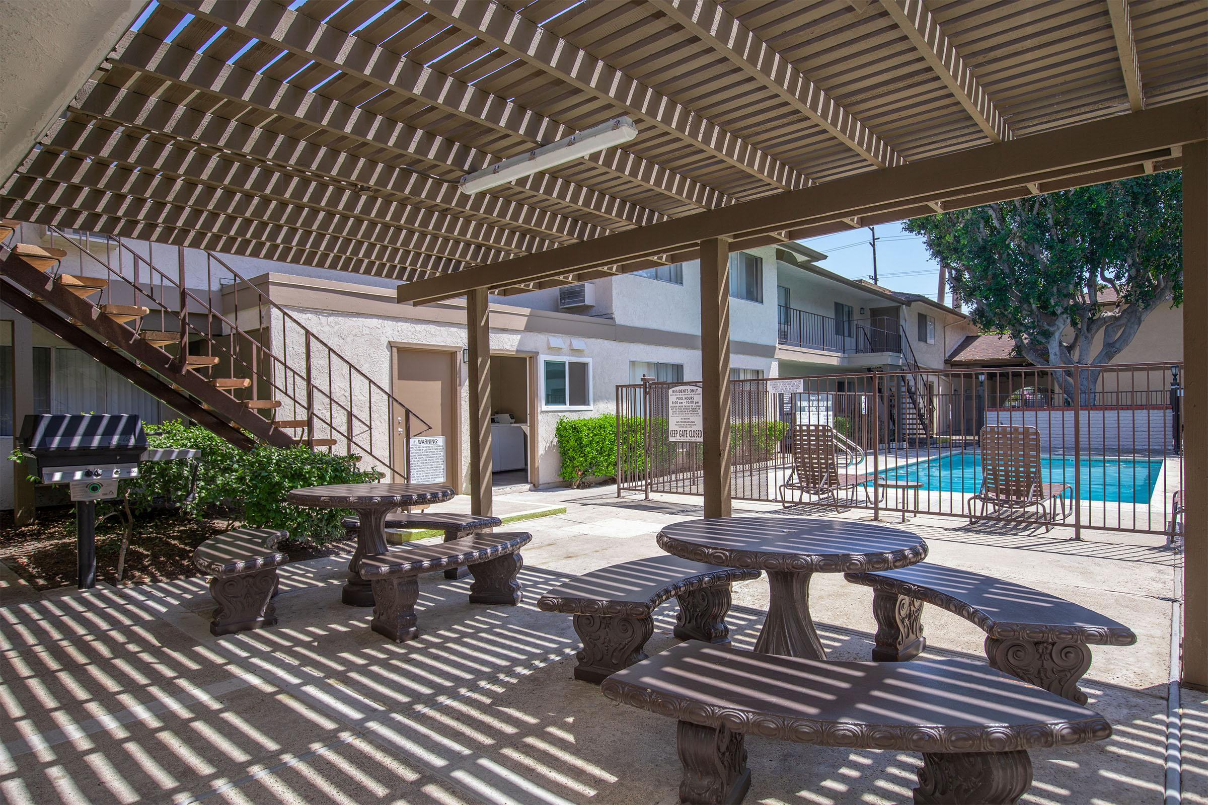 Outdoor area featuring picnic tables under a pergola, with a swimming pool in the background. There are fences around the pool, a grill nearby, and a staircase leading to upper levels of the building. Sunlight creates a pattern through the slats of the pergola.