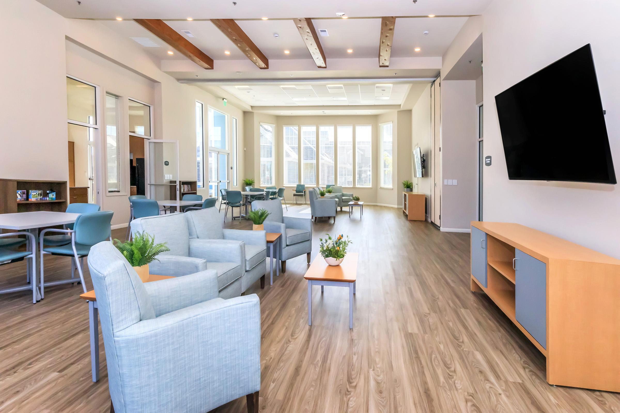 A spacious lounge area featuring light-colored walls and wooden beams. The room includes comfortable seating with soft blue chairs, small tables, and a large TV mounted on the wall. Abundant natural light floods in through large windows, creating a welcoming atmosphere.