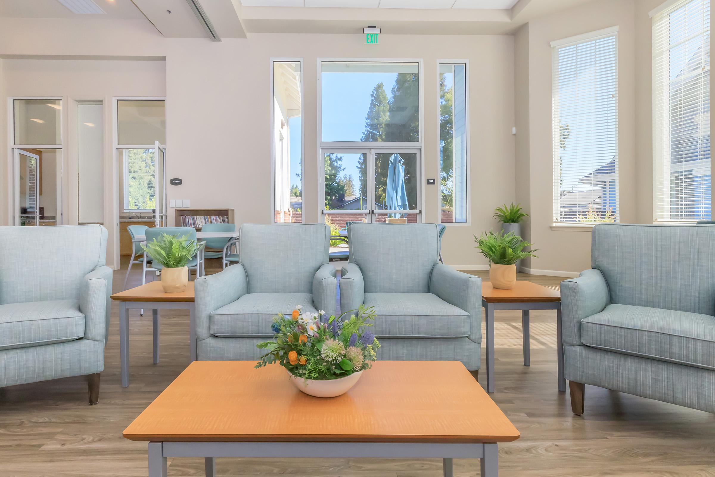 A modern lounge area featuring light blue upholstered chairs with small wooden side tables. A coffee table in the center holds a decorative arrangement of flowers. Large windows allow natural light, with green plants visible outside. The space is bright, airy, and inviting, ideal for relaxation or conversation.
