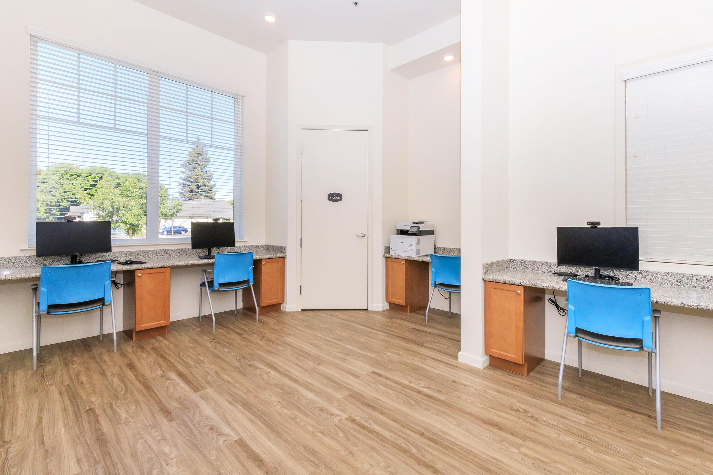 A bright, modern workspace featuring several desks with computers, blue chairs, and a printer. Large windows allow natural light to fill the room, which has light-colored walls and wooden flooring. The atmosphere is clean and inviting, ideal for productivity.