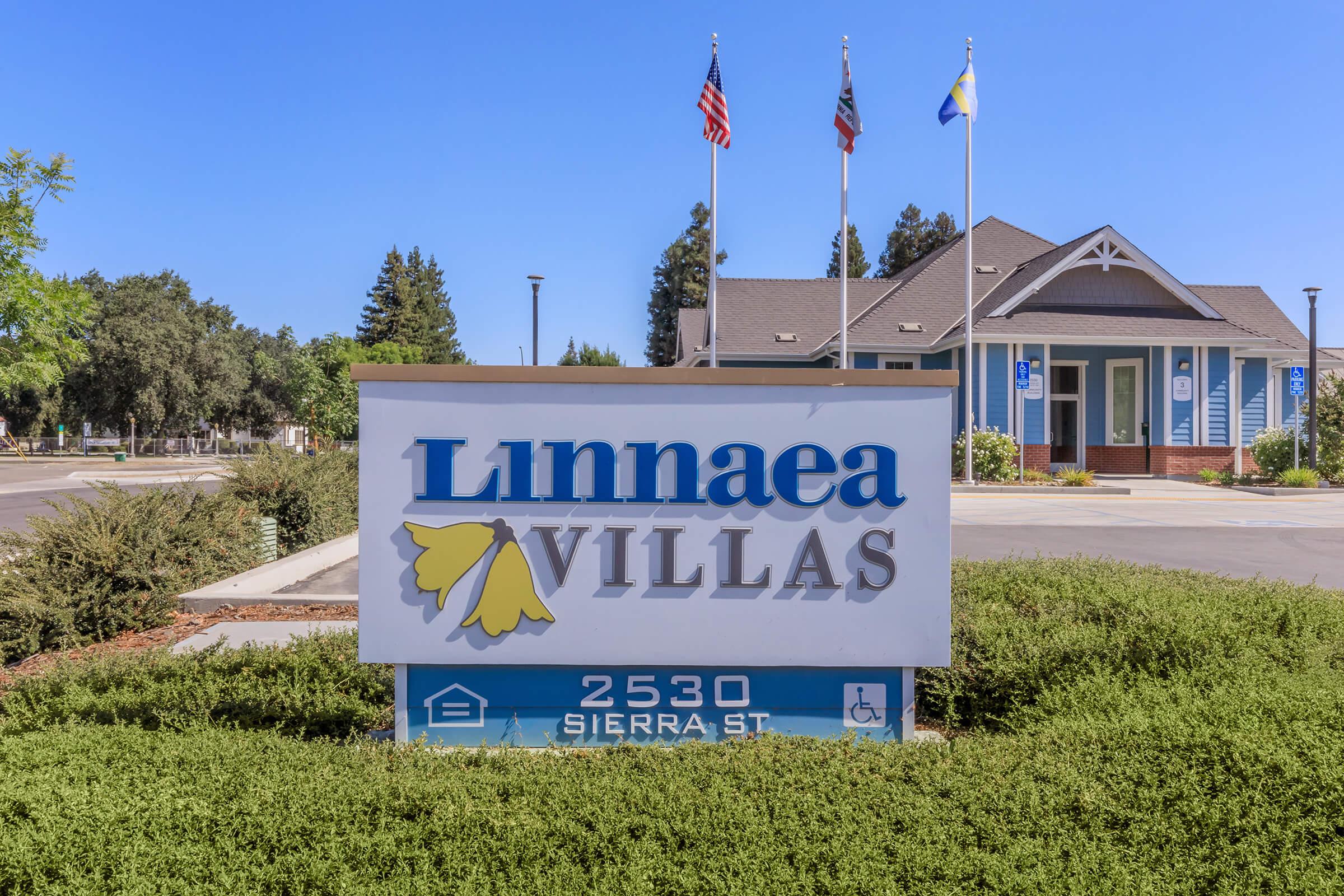 Sign for Linnaea Villas located at 2530 Sierra St, featuring the name prominently displayed alongside decorative elements and landscaping. Flags are visible in the background, with a residential building nearby.