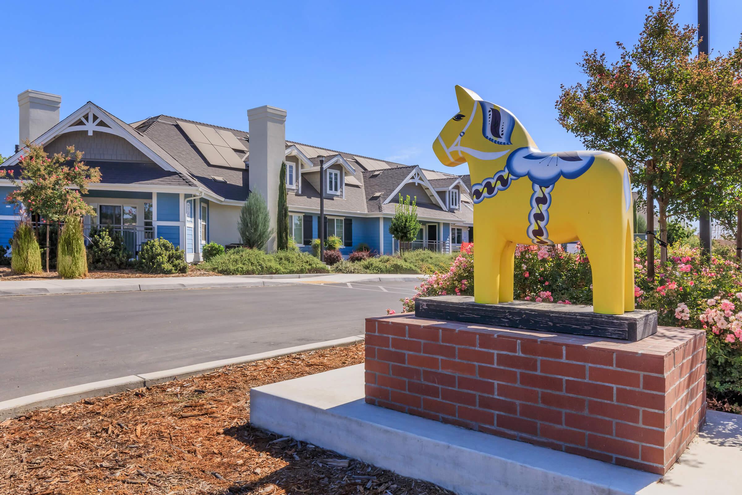 A bright yellow wooden horse statue with a blue and white design stands on a brick base in front of a residential building. The building features a mix of blue siding and white trim, surrounded by well-maintained greenery and flowers under a clear blue sky.