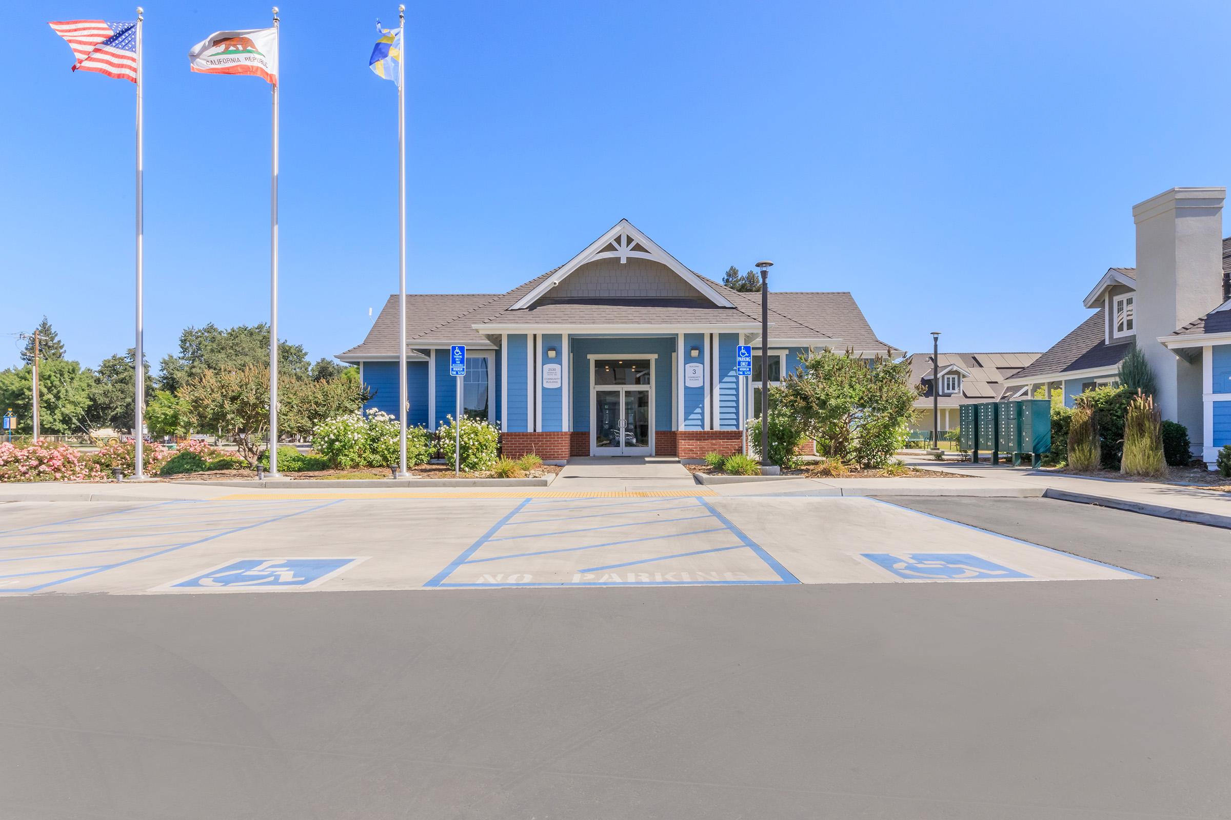 A blue building with a distinctive architectural style, featuring a front entrance and surrounded by landscaped greenery. Three flags are flying in front, with designated handicap parking spaces visible in the foreground. The sky is clear and blue, indicating a bright day.