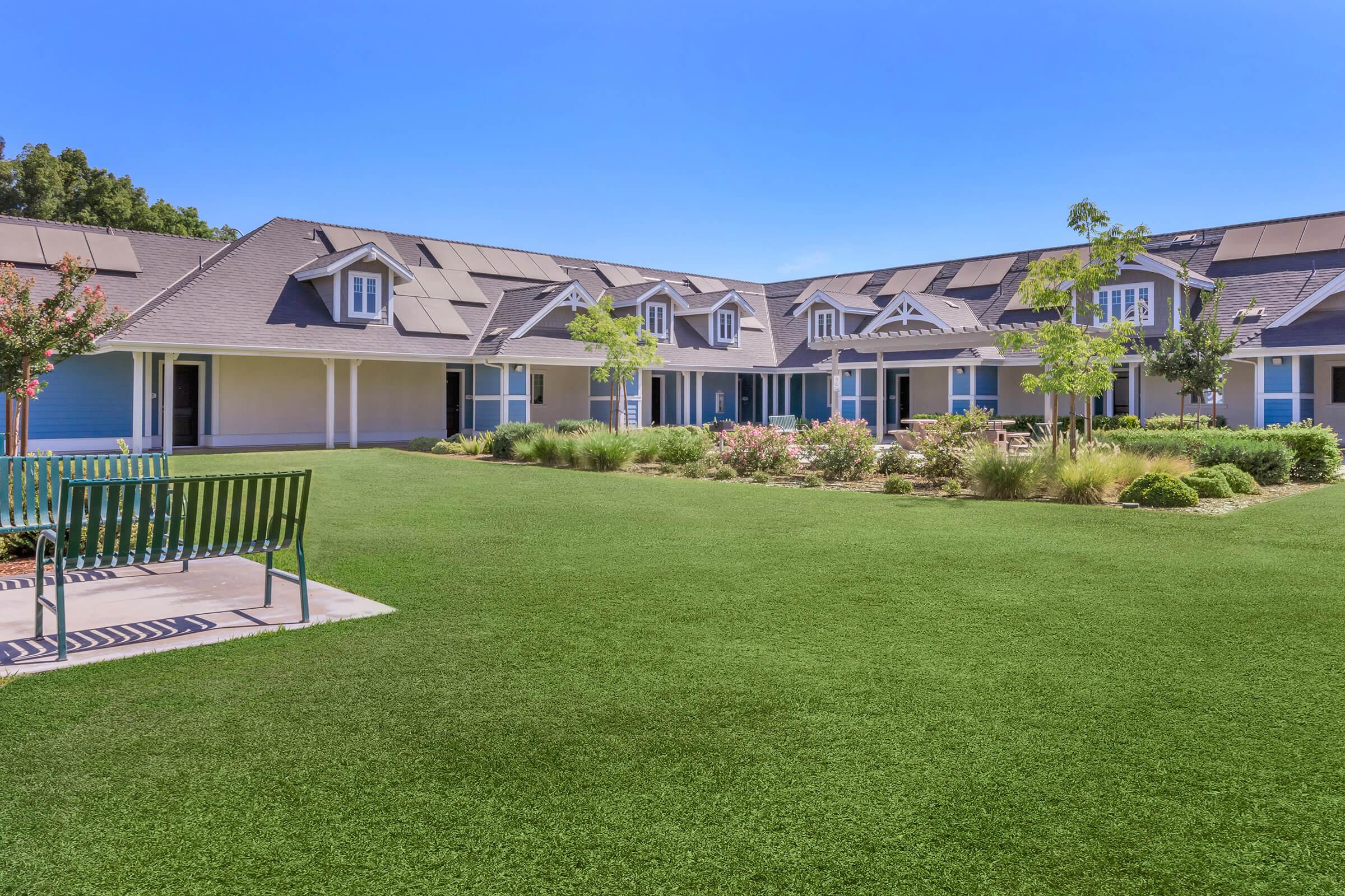 A landscaped courtyard with well-maintained green grass, surrounded by buildings featuring gray roofs and blue siding. Flowering plants and small trees enhance the garden area, while a bench offers a seating space amid the serene environment. Clear blue sky overhead.