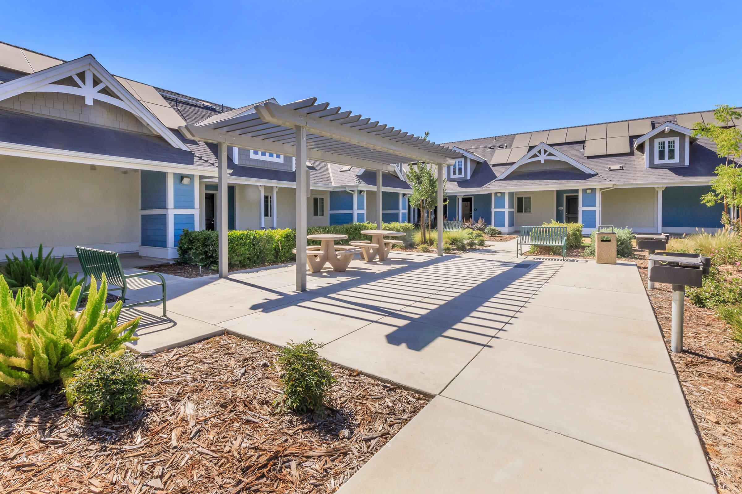 A sunny courtyard featuring a pergola casting shadows on a concrete path. Surrounding the area are well-maintained shrubs and benches. The background shows a series of residential buildings with a modern aesthetic, showcasing a clear blue sky.
