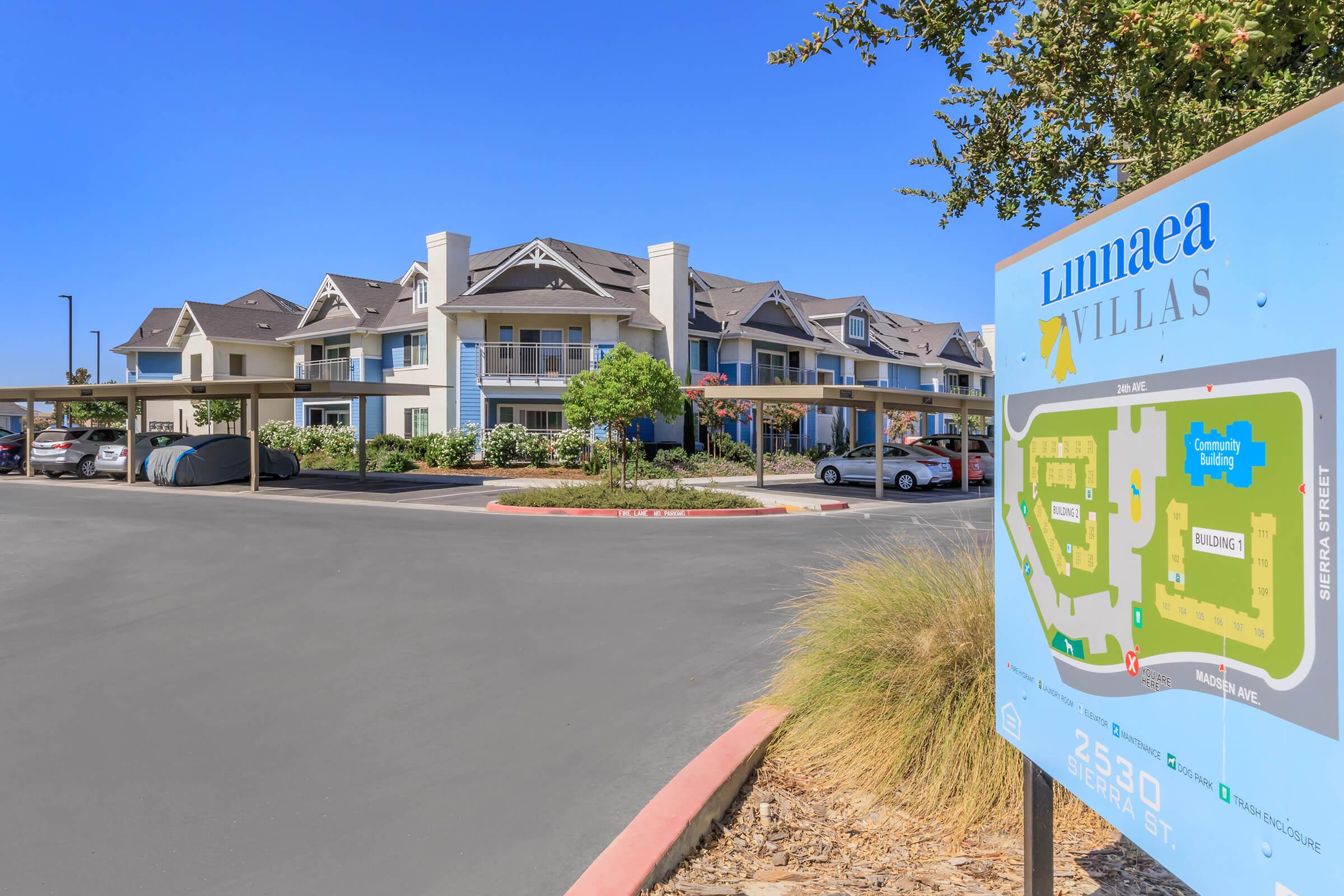 A parking area leading to Luminea Villas, featuring a multi-story building with a mix of blue and beige exterior. A sign displays a map of the property, indicating the location of the community building, as well as designated parking and landscaped areas. Trees and cars are visible in the foreground.