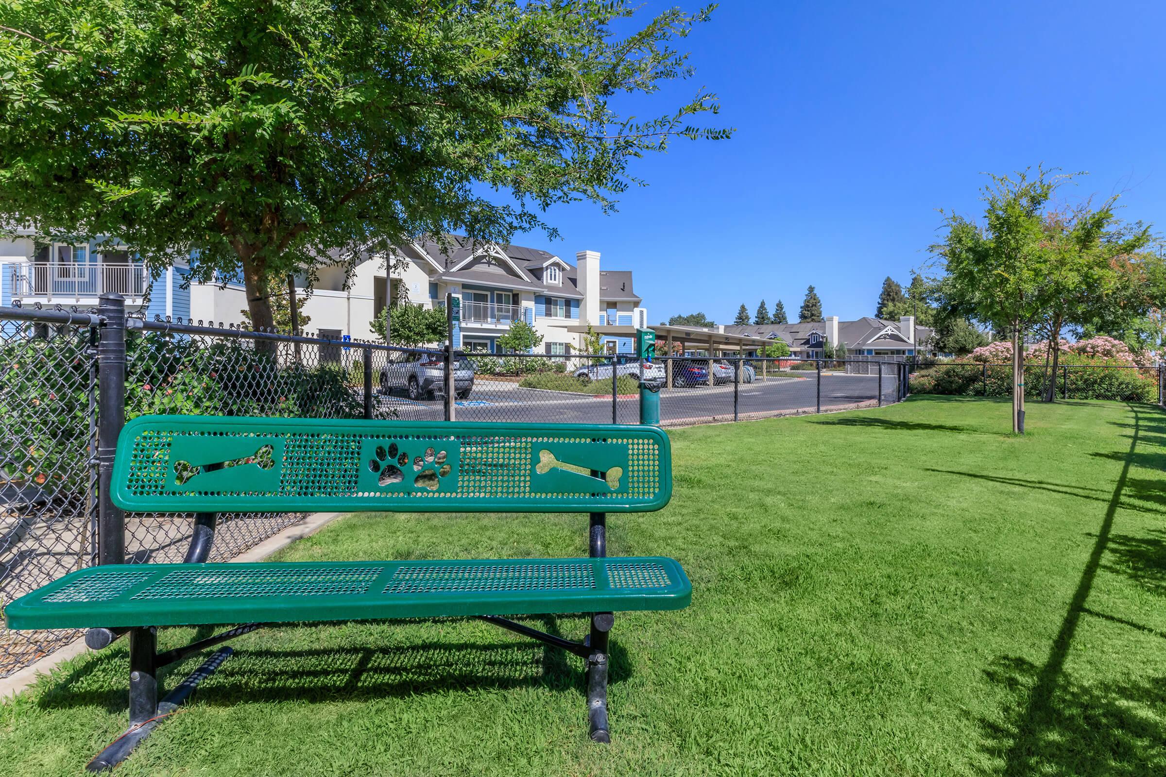 A green park bench featuring dog bone and paw print designs, set on a well-maintained grassy area. In the background, a gated community with homes and parked cars is visible under a clear blue sky. Trees provide some shade in the area.