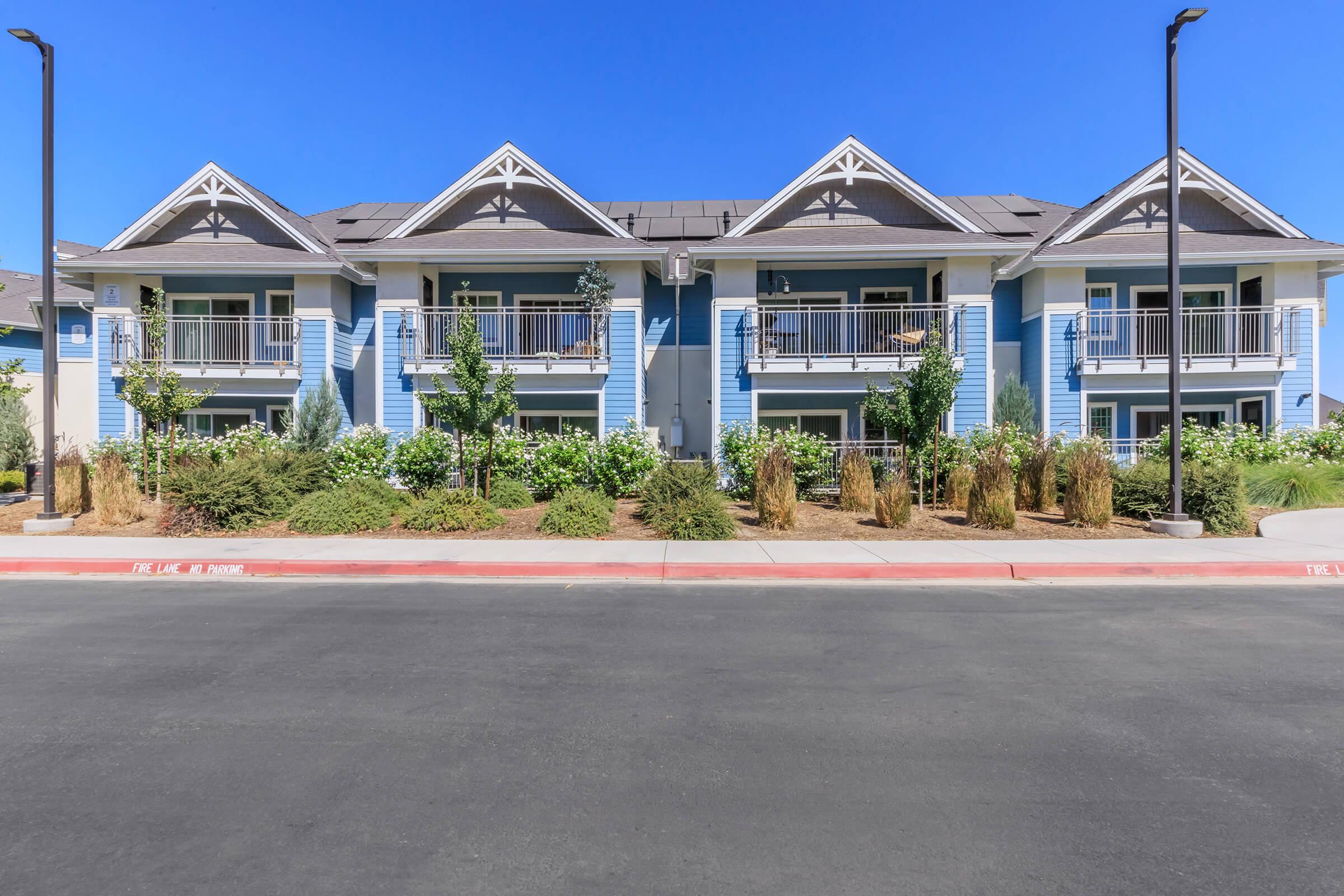 A two-story residential building with a light blue exterior, featuring multiple balconies with railings. The landscaping includes shrubs and decorative plants, with a clear blue sky above. The road in front is well-paved, suggesting a modern and well-maintained living environment.