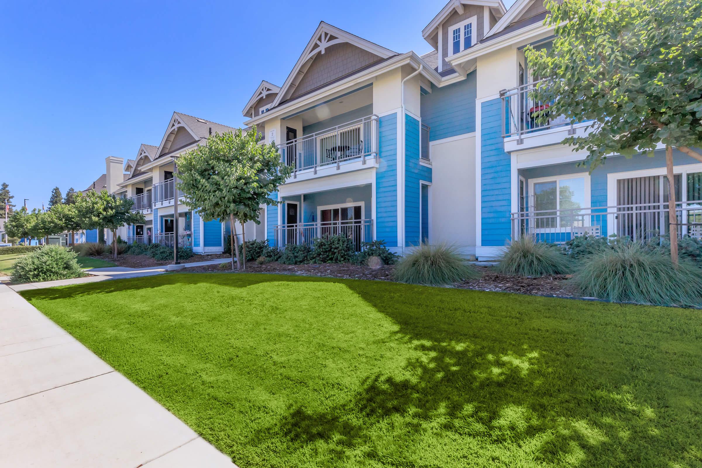 A modern apartment complex with blue and white exterior. The building features balconies and is surrounded by manicured lawns and small trees. The sidewalk runs alongside the grassy area, creating a welcoming outdoor space. Clear blue sky overhead.