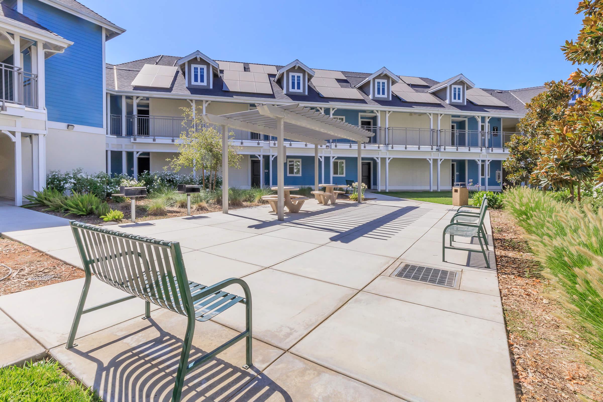 A courtyard with green benches and a pergola is surrounded by buildings. The area features well-maintained grass and landscaping, with pathways leading to various seating arrangements. The sky is clear and sunny, creating a bright and inviting atmosphere.