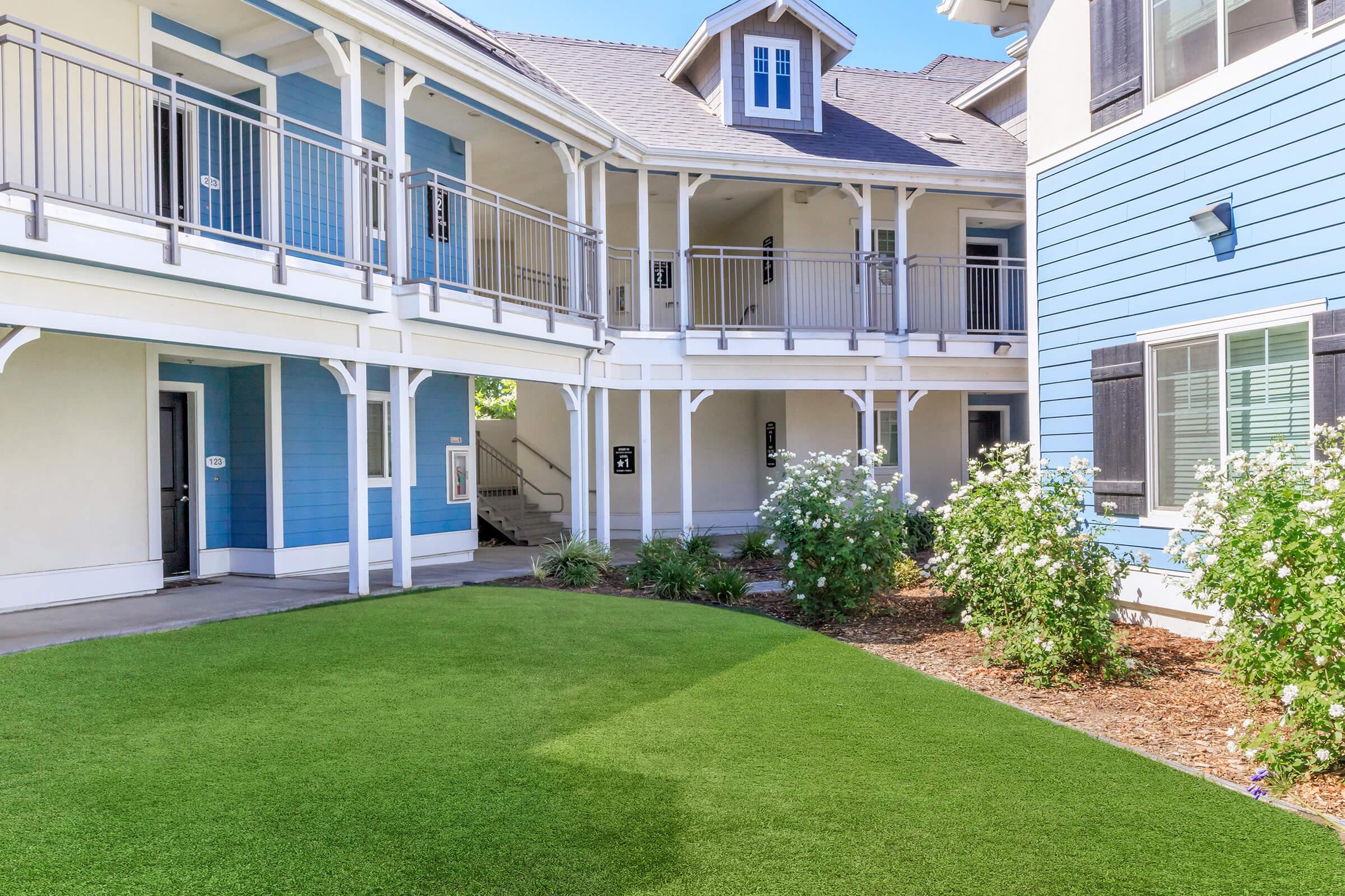 A well-maintained courtyard with lush green artificial grass, surrounded by buildings featuring a mix of blue and white exteriors. Flowering shrubs add color to the space, while balconies provide access to the upper levels of the buildings. Bright sunlight illuminates the area, creating a welcoming atmosphere.