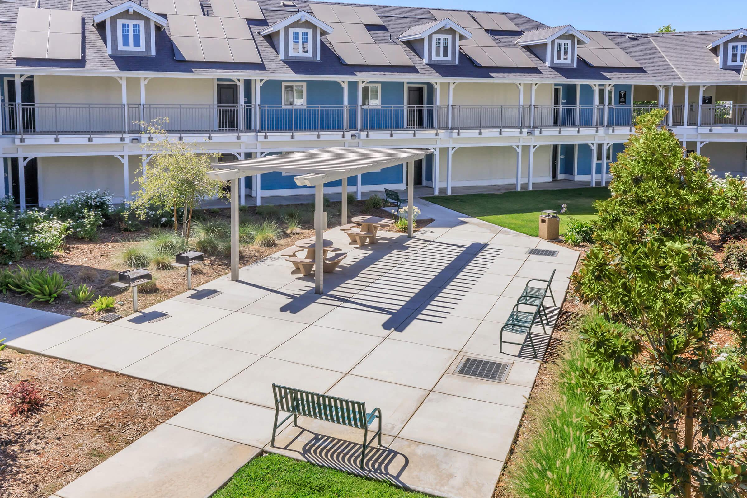 A view of a landscaped courtyard featuring a concrete patio with picnic tables and benches, surrounded by greenery. The background shows a building with multiple residential units and solar panels on the roof, enhancing sustainability and inviting communal space.