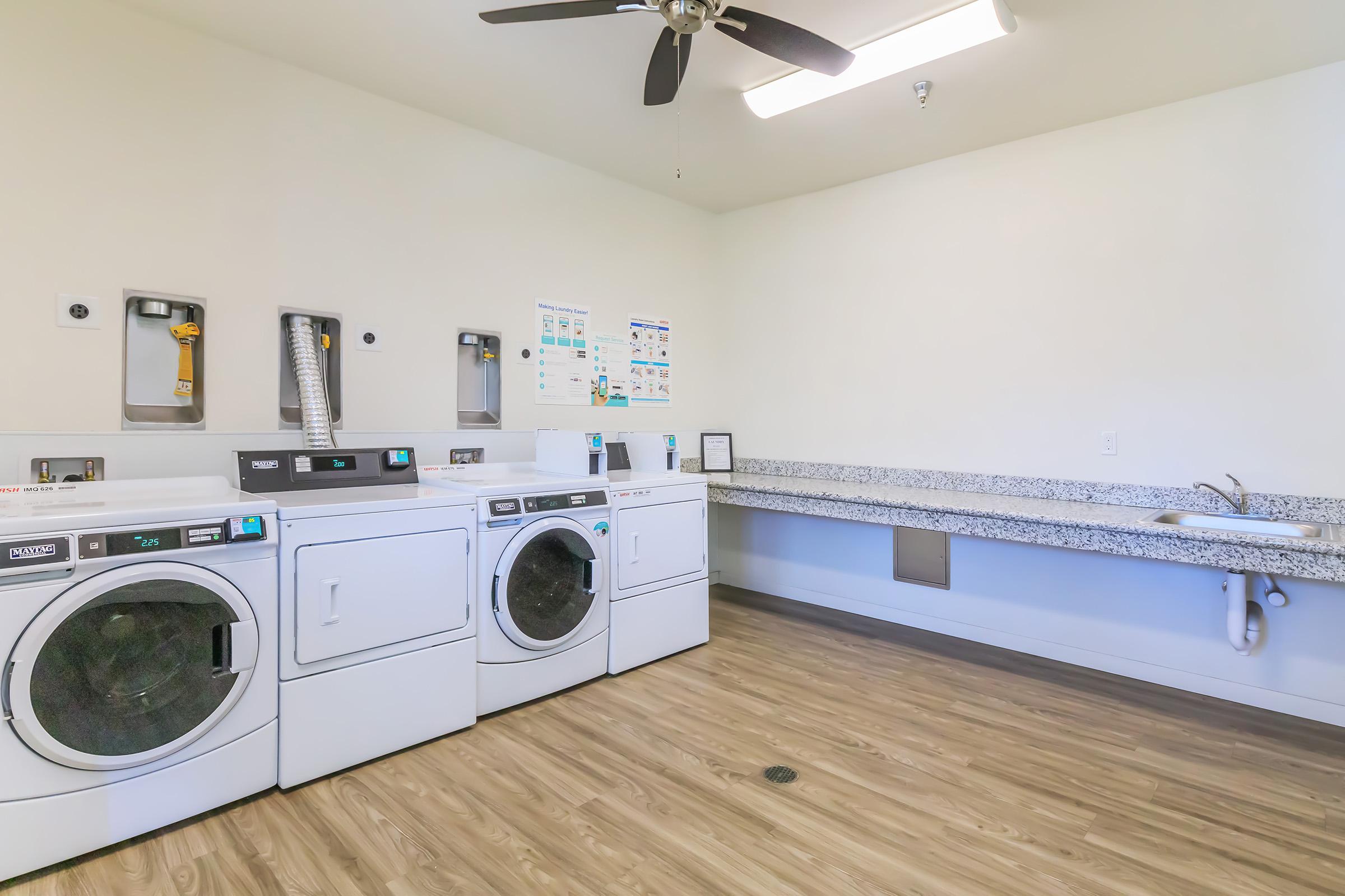 A clean, bright laundry room featuring three white washing machines and dryers, a long counter with a sink, and a ceiling fan. The walls are painted a light color, and there are informational posters about laundry care. The floor is made of faux wood planks.