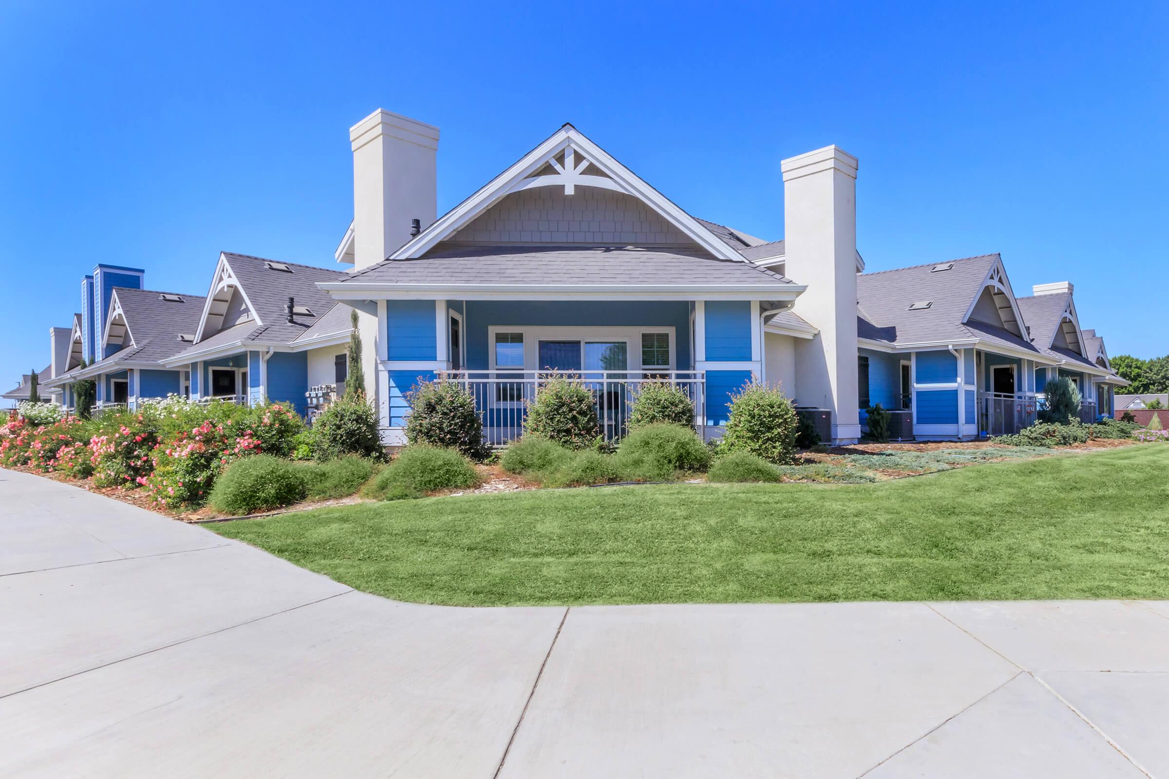 A modern residential building with blue and beige accents, featuring a well-manicured lawn in the foreground. The structure has multiple chimneys, large windows, and a covered porch. Bright blue sky overhead complements the inviting atmosphere of the property.