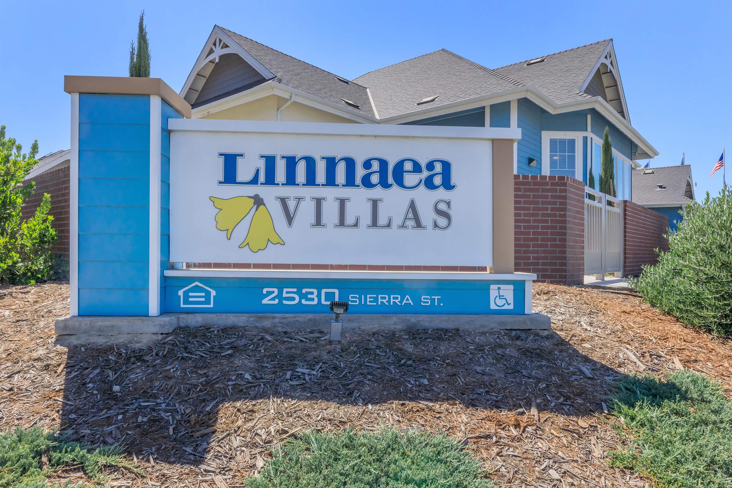 Sign for Linnaea Villas located at 2530 Sierra St. The sign features a blue and white color scheme with a yellow flower graphic, surrounded by landscaped areas. In the background, there are trees and a building with a gabled roof.