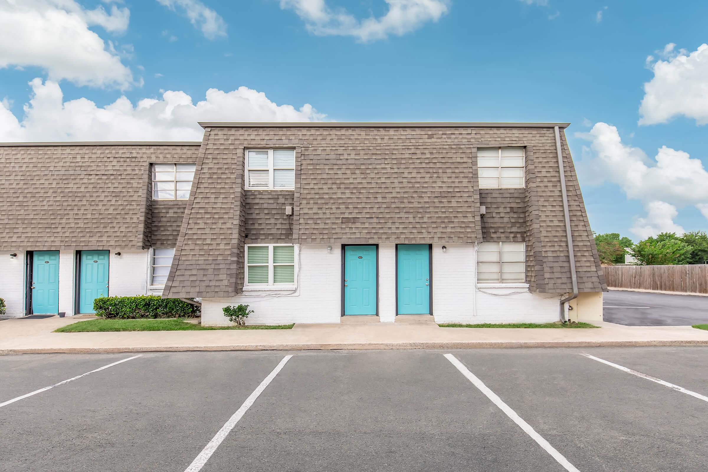 A two-story apartment building featuring a sloped roof, with multiple units each having a turquoise door. The building is surrounded by green shrubbery and has a parking lot in front, displaying several empty parking spaces. The sky is bright with fluffy clouds.