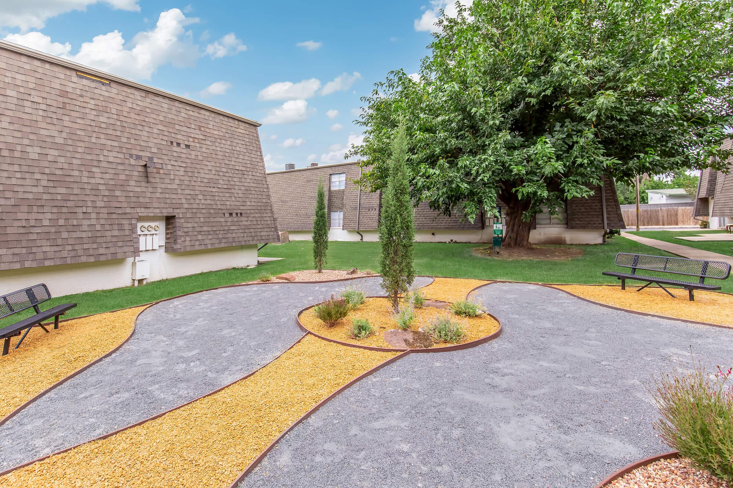 A landscaped courtyard featuring two winding pathways made of gravel and bordered by yellow pebbles. In the center, there is a circular garden area with small shrubs and young trees. Benches are placed along the pathways, and surrounding buildings have a rustic, wooden exterior under a blue sky with scattered clouds.