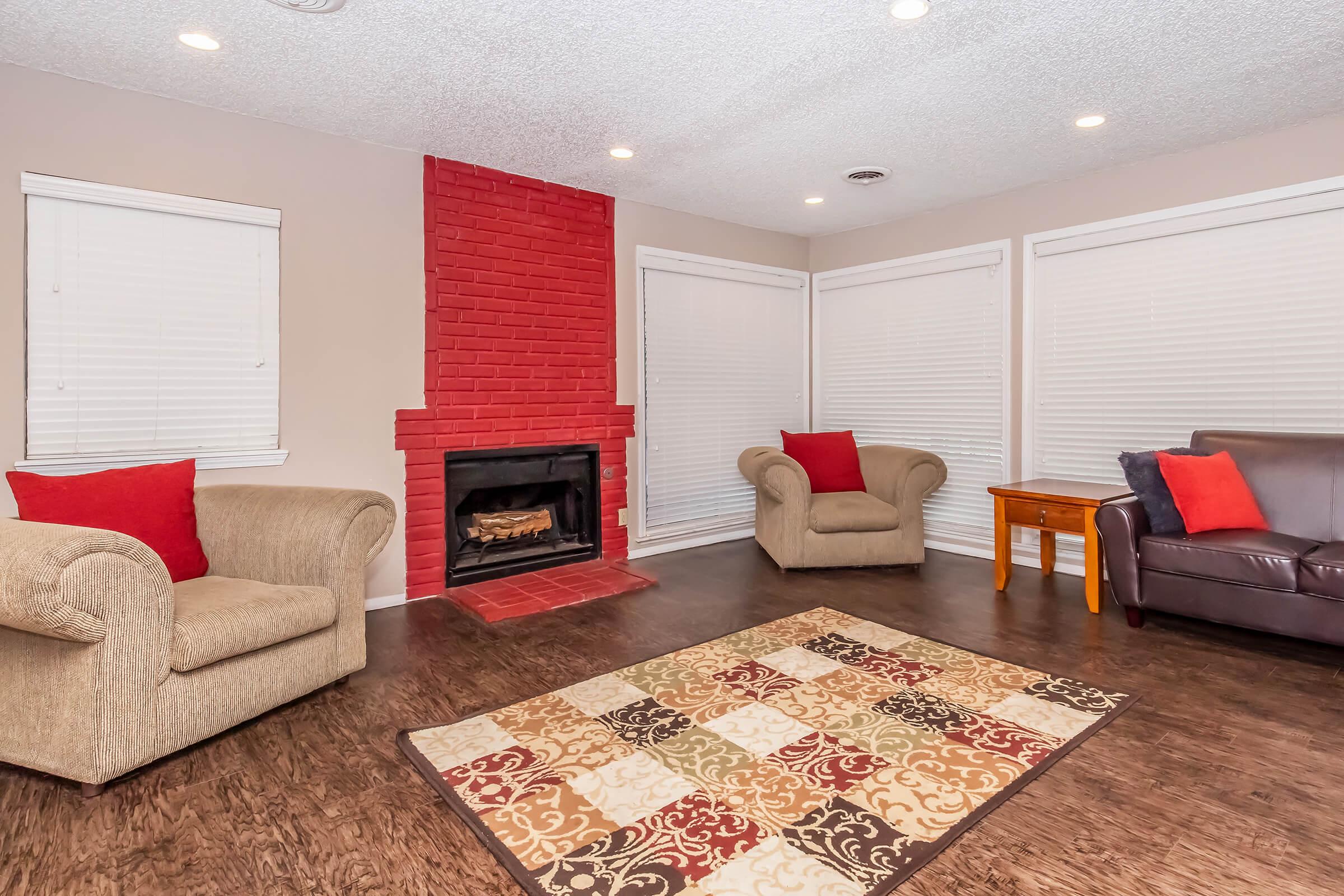 A cozy living room featuring a red brick fireplace, two armchairs with red pillows, and a small wooden side table. The floor is covered with a patterned area rug, and there are white window blinds in the background, creating a warm and inviting atmosphere.
