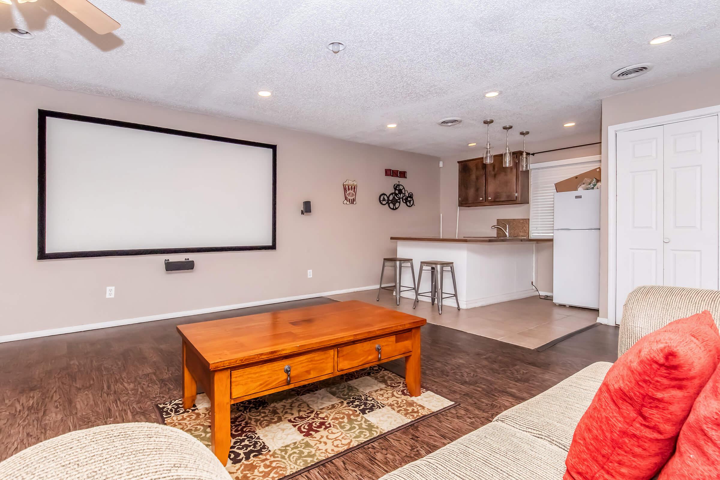 A spacious living room featuring a large blank screen on the wall, a wooden coffee table, and a sofa with red cushions. In the background, there's a kitchen area with barstool seating and a refrigerator, all under soft lighting and a textured ceiling.