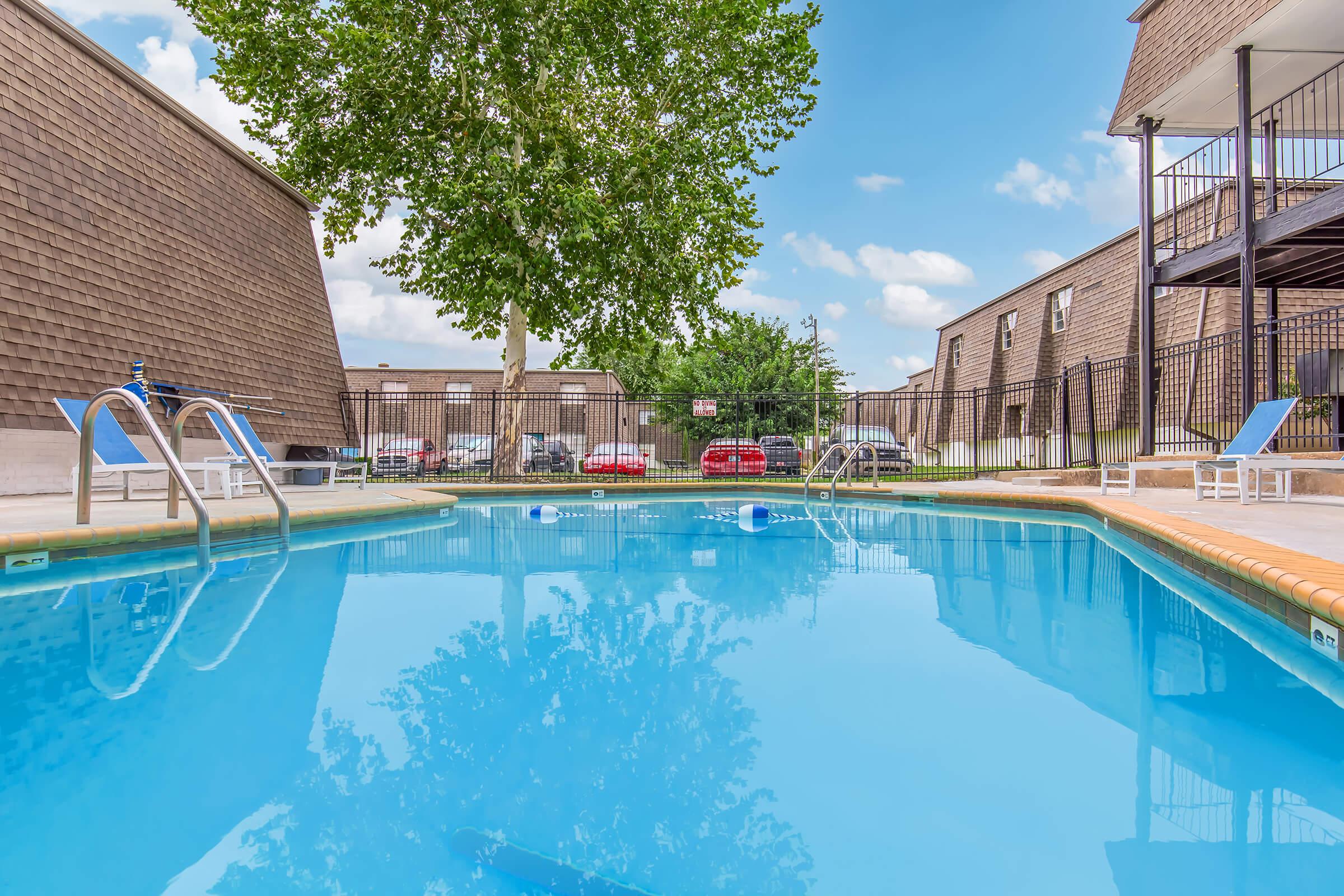 A clear blue swimming pool surrounded by a fence, with lounge chairs on the deck. Trees and apartment buildings are visible in the background under a bright blue sky with fluffy clouds. Cars are parked nearby, adding to the residential setting.