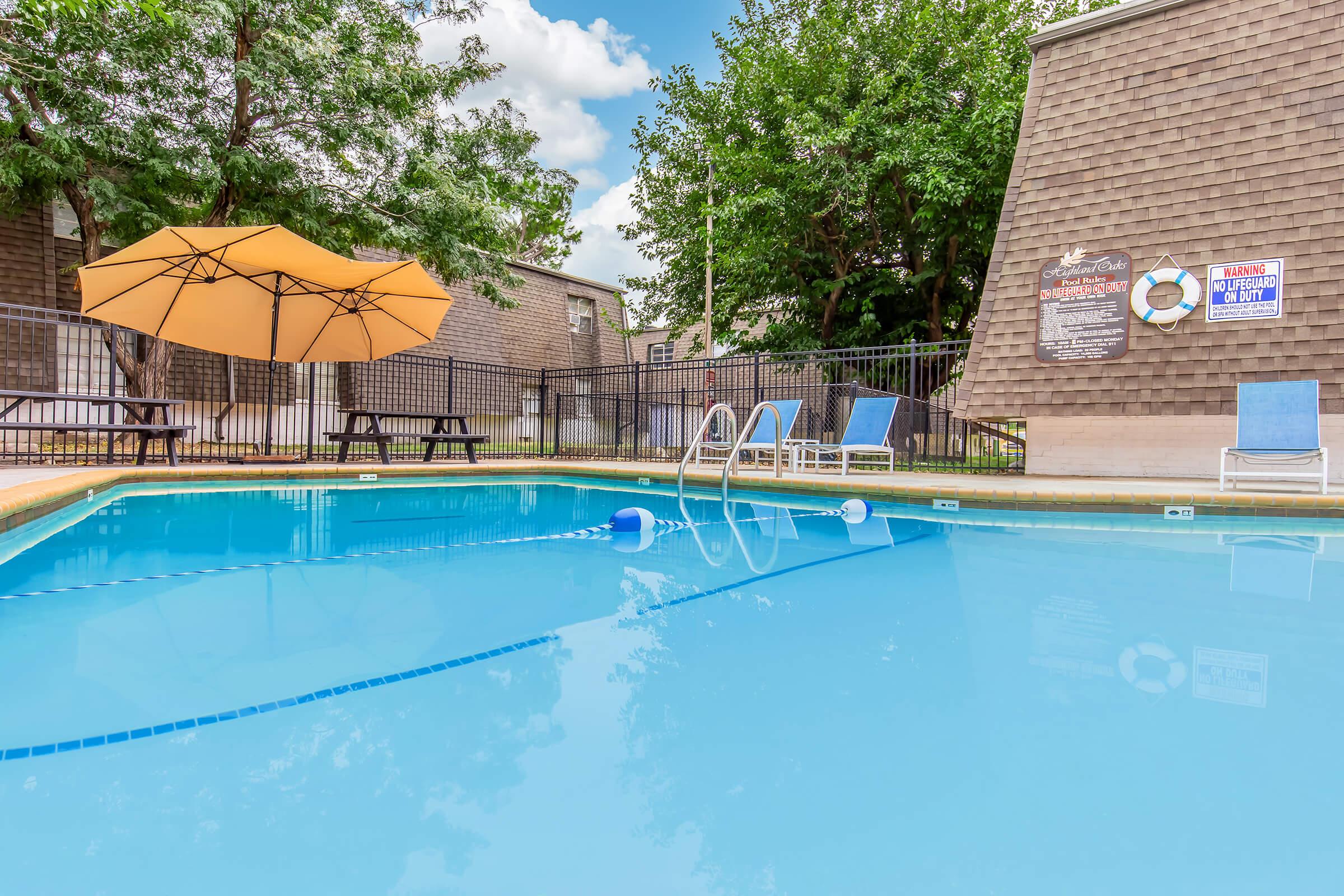 A clear, inviting swimming pool surrounded by green trees. There are two blue lounge chairs by the pool, an orange umbrella providing shade, and a few picnic tables nearby. The pool features a diving board and is surrounded by a black fence. In the background, there are buildings with a textured exterior.