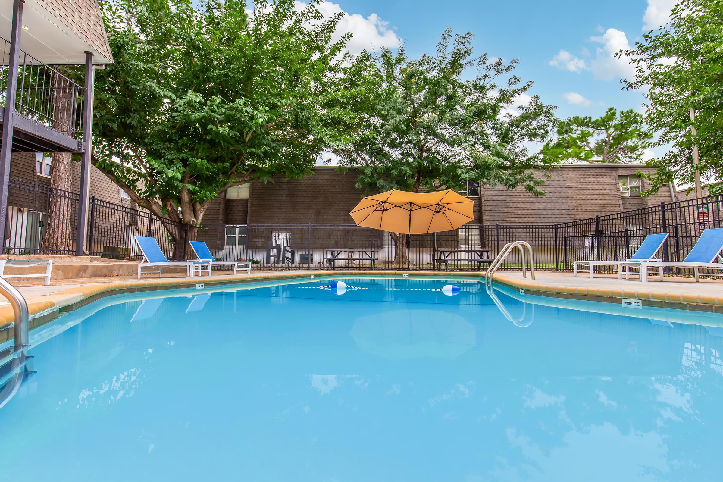 A clear swimming pool surrounded by lounge chairs and an umbrella, with green trees in the background and a blue sky above. The pool area is clean and inviting, perfect for relaxation and leisure activities.