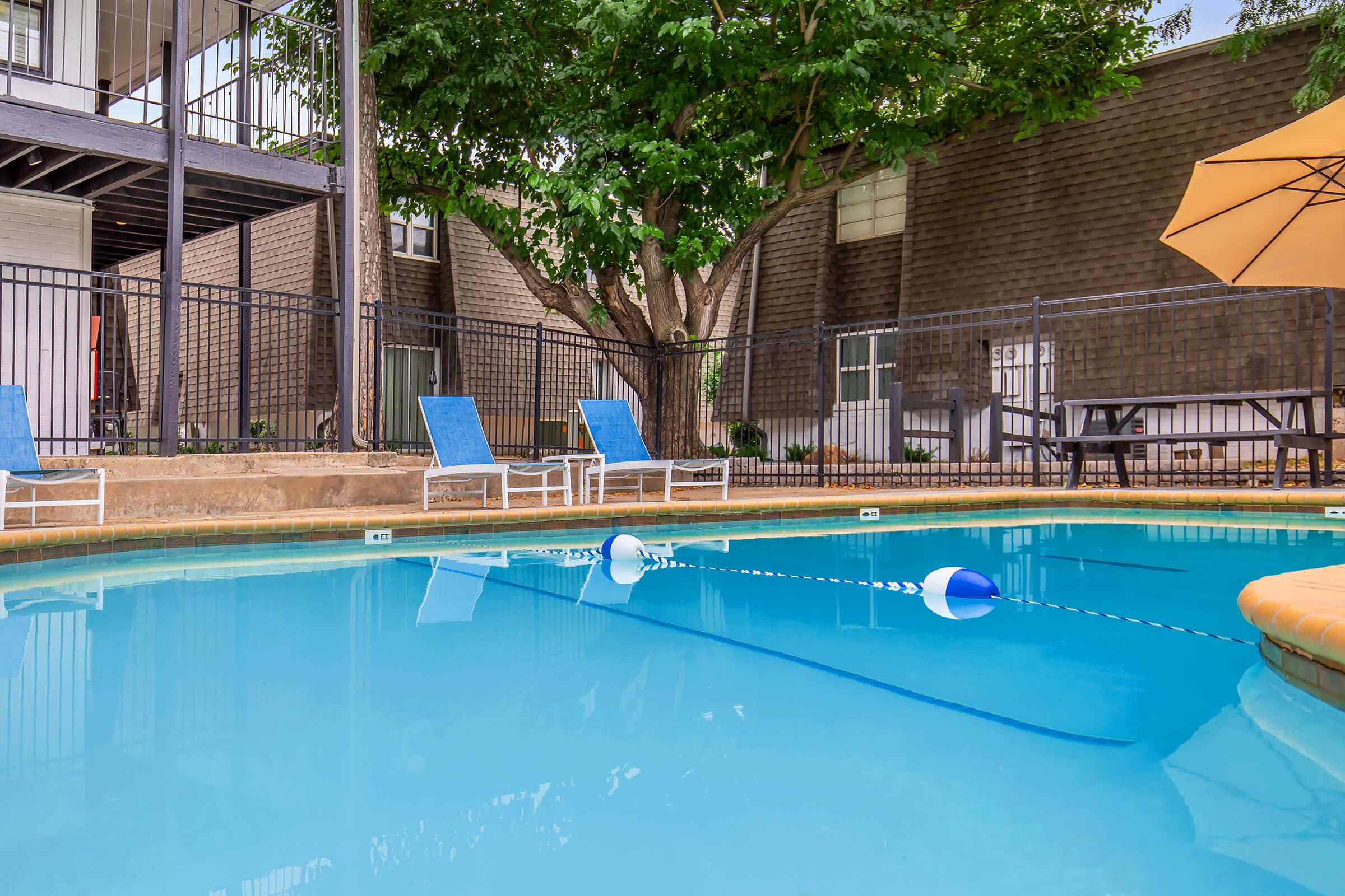 A swimming pool with clear blue water, bordered by a tan tile deck. There are two blue lounge chairs on one side and a yellow umbrella nearby. A buoy line is visible in the water, and there's a shaded area with trees and a fence surrounding the pool area.