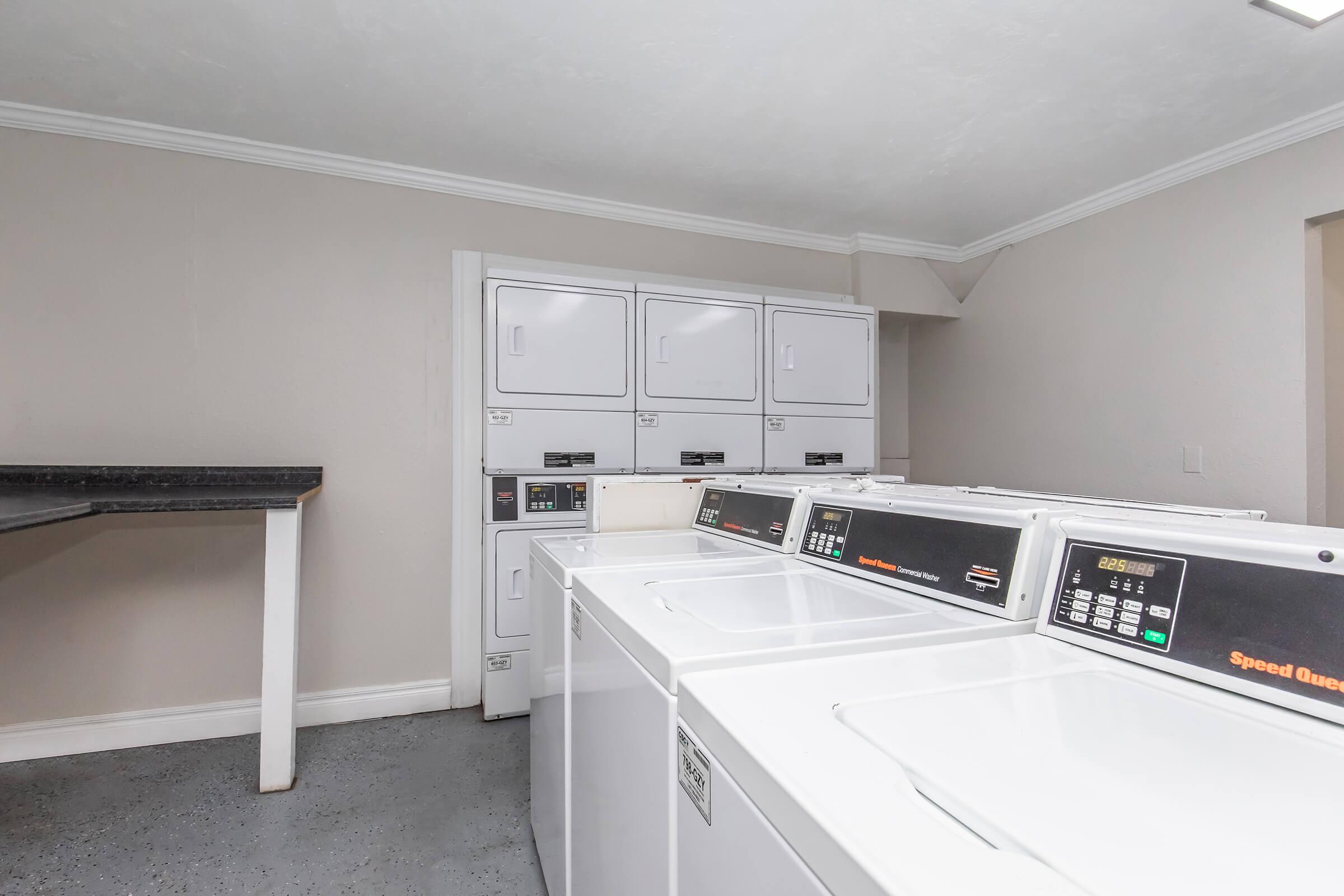 A clean laundry room featuring several white washing machines and dryer units. The machines have digital displays and are arranged neatly. There is a black countertop against the wall for folding clothes, with light-colored walls and a well-lit ambiance, creating a tidy and organized space for laundry activities.