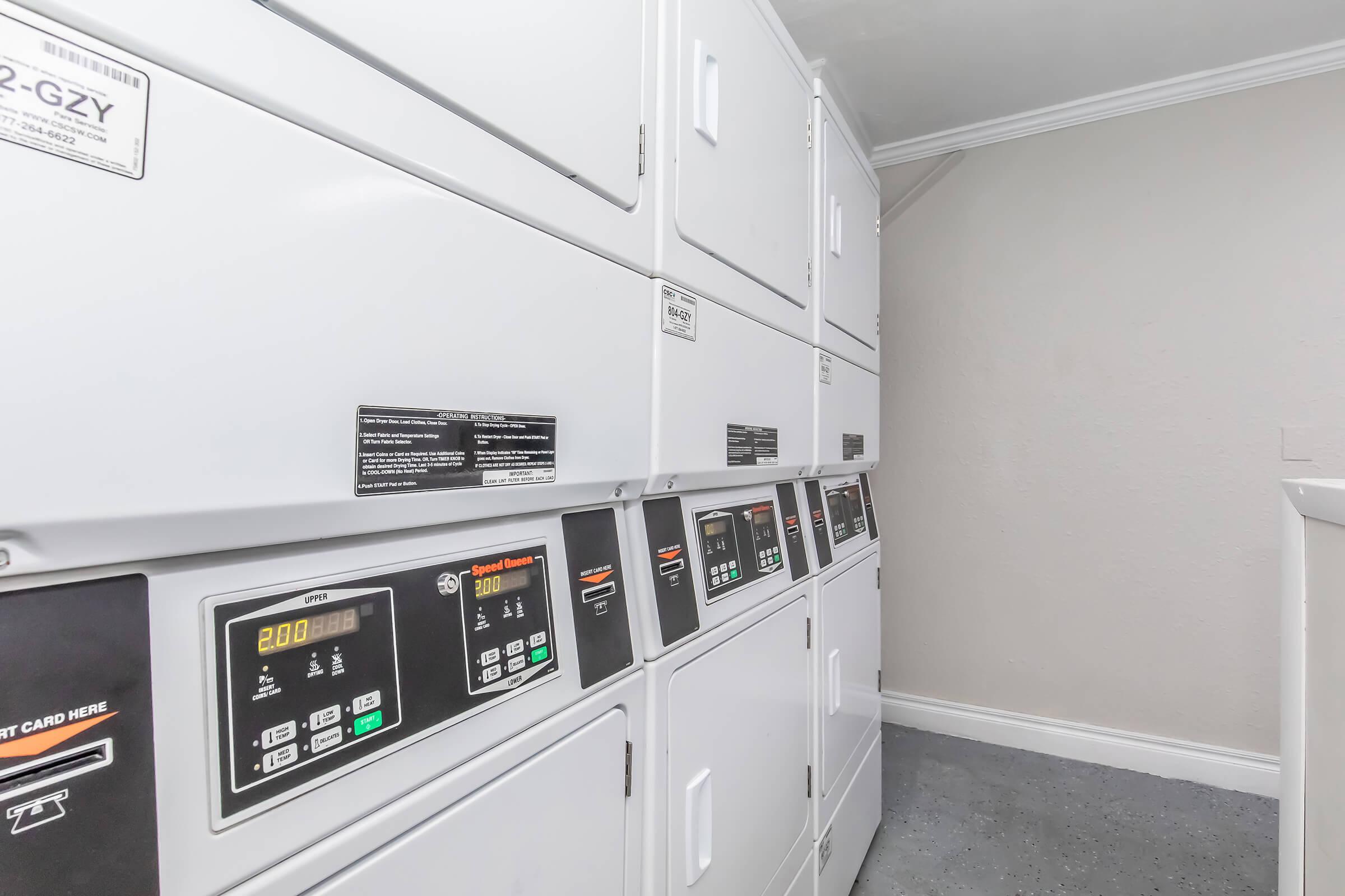 A row of white laundry machines in a clean laundry room, featuring digital displays and card readers. The machines are neatly arranged with labels indicating different settings and prices for wash cycles. The room has a light-colored wall and a simple, functional design.