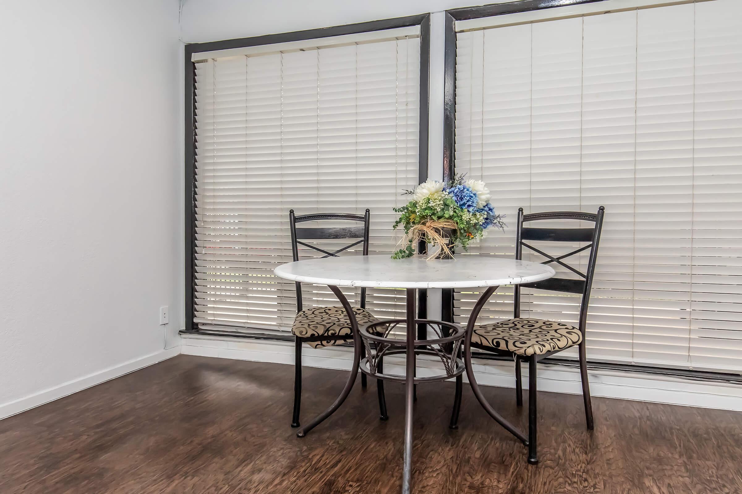 A small dining area featuring a round marble table with two black metal chairs, one with a patterned cushion. A floral arrangement in a vase is centered on the table. Large windows with blinds provide natural light, and the floor is a dark wooden finish.