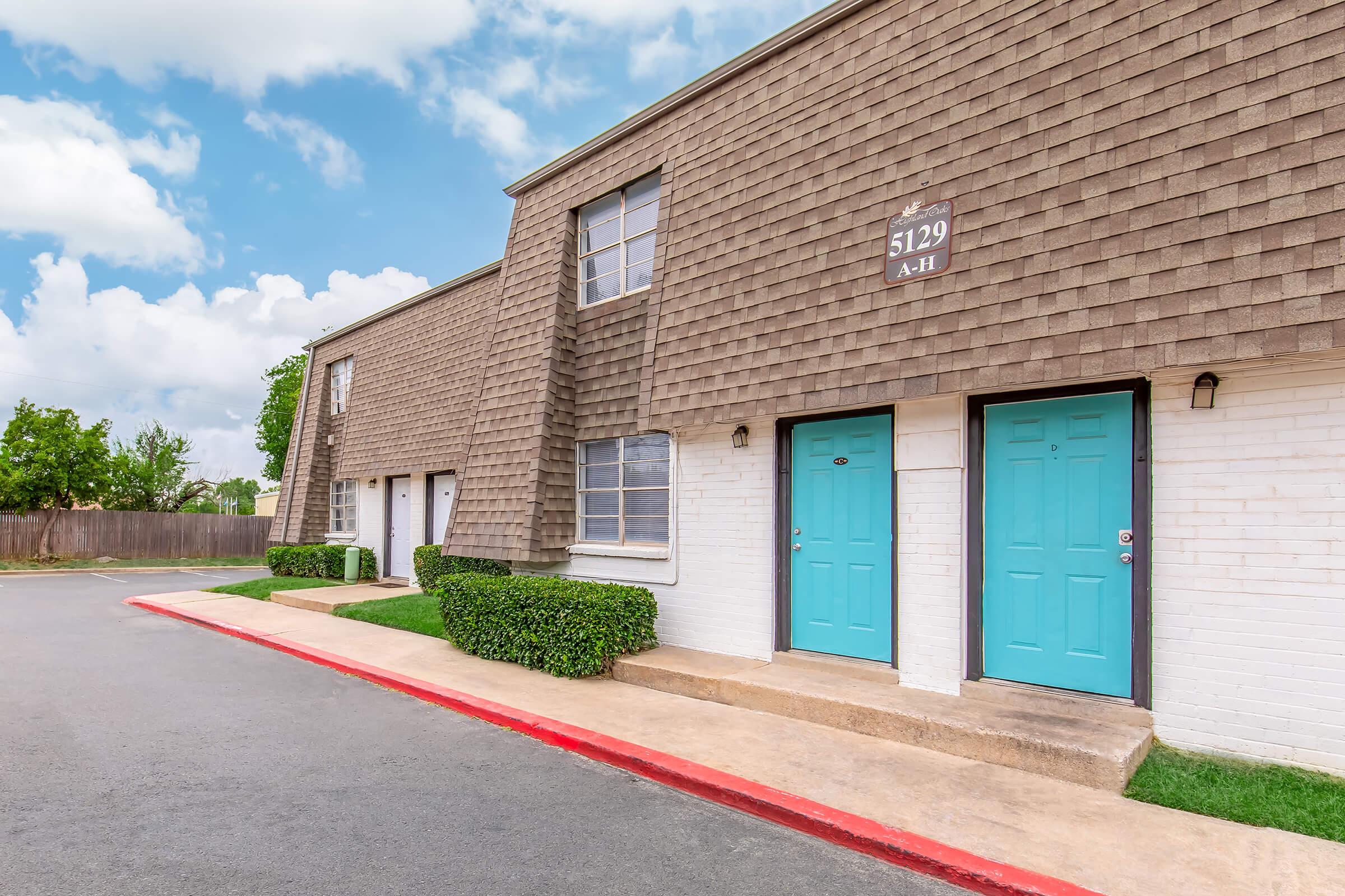 Two-story apartment building with a textured brown exterior. The building features several doors painted in bright turquoise, each with a numbered plaque above. A landscaped area with small bushes lines the walkway, and a clear blue sky with some clouds is visible in the background.