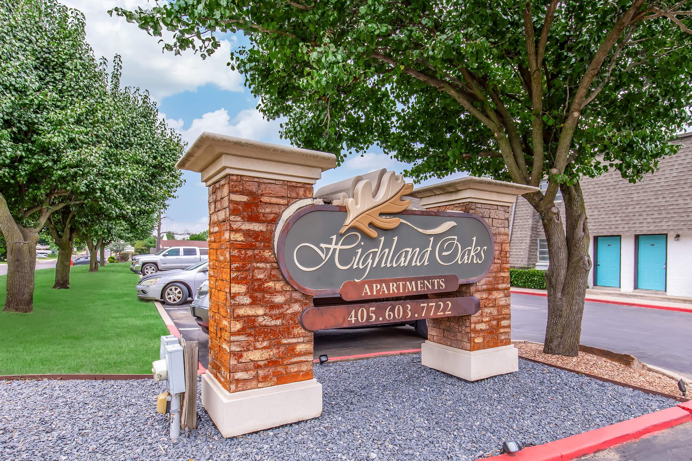 Sign for Highland Oaks Apartments featuring an elegant design with a leaf motif. The sign includes the name "Highland Oaks Apartments" and a phone number, set against a backdrop of trees and landscaped grounds. Nearby vehicles and buildings are visible, with a clear sky in the background.